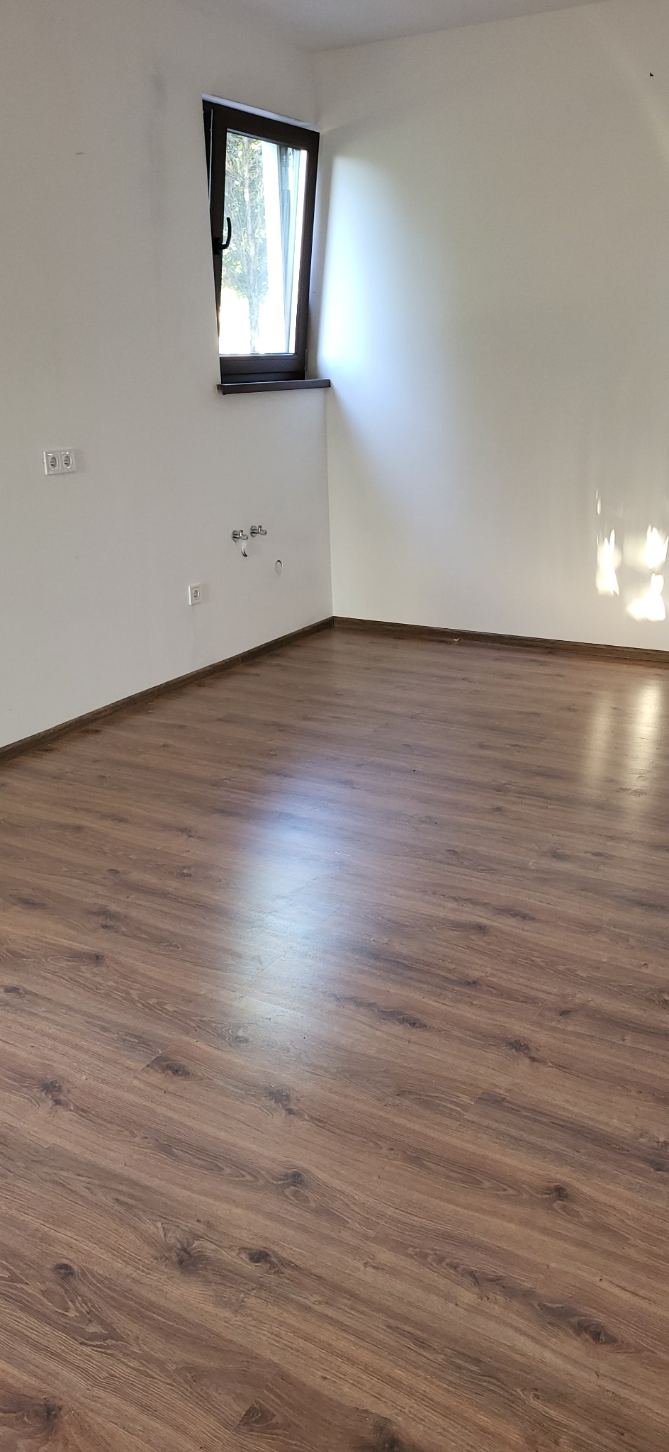 Puste, jasne wnętrze po remoncie: brązowe panele podłogowe, białe ściany z gniazdkami i instalacją hydrauliczną, okno z widokiem na zewnątrz.