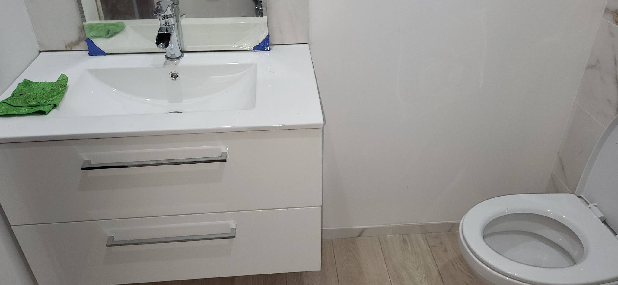 Biała umywalka z szafką w nowoczesnej łazience, widoczny kran, lustro z folią ochronną i toaleta. Świeżo po remoncie, czysta przestrzeń.