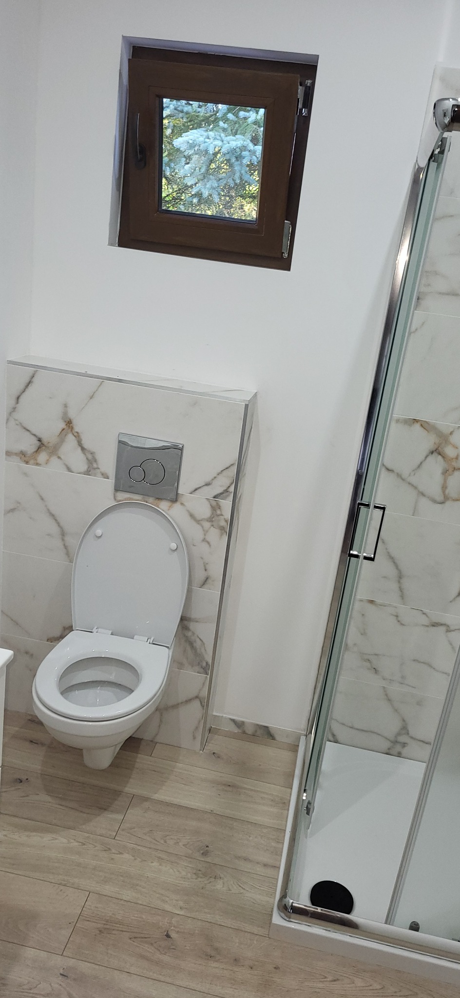 Wykończona łazienka: toaleta z marmurową obudową, kabina prysznicowa z marmurowymi płytkami i drewnopodobna podłoga. W tle okno z widokiem na drzewo.