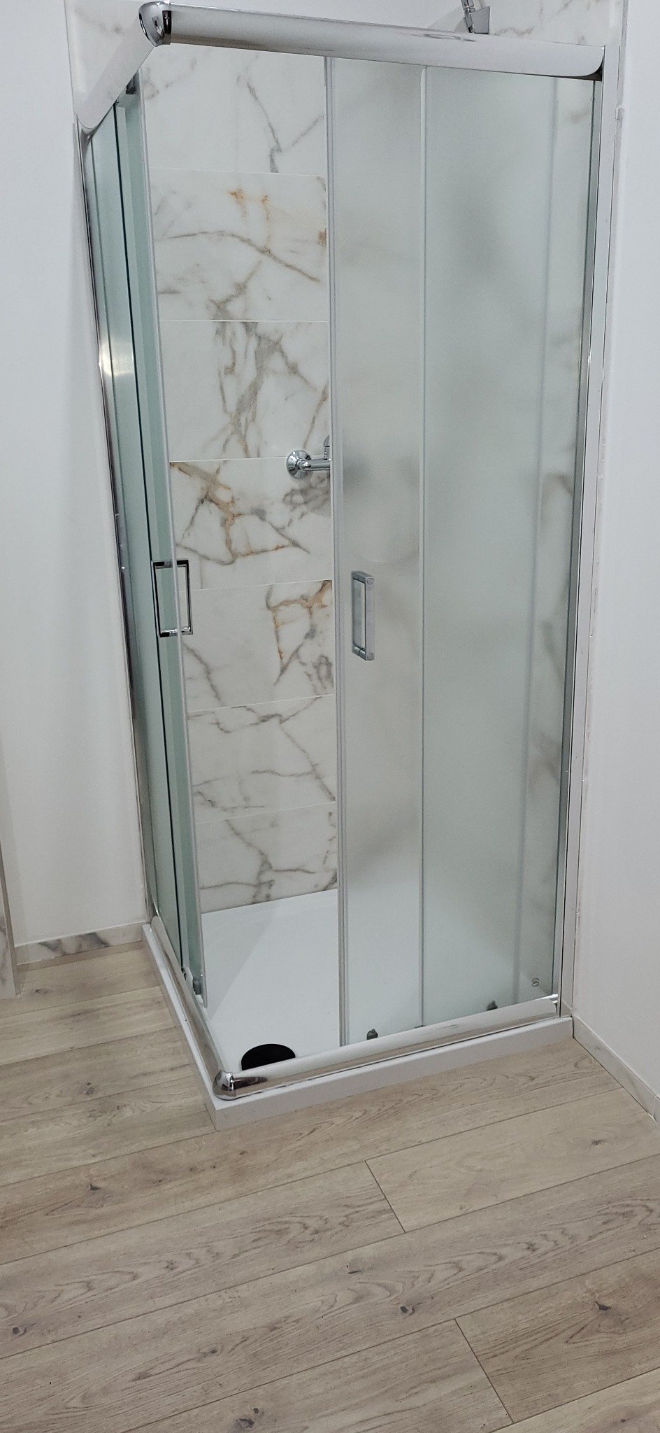 Nowoczesna kabina prysznicowa narożna z przesuwnymi drzwiami, wykończona chromowanymi profilami. Wnętrze wyłożone płytkami imitującymi marmur. Jasna podłoga.