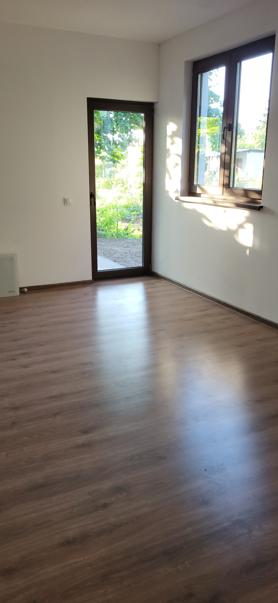 Pusty pokój z drewnianą podłogą, oknem z widokiem na zieleń i brązowymi ramami. Świeżo pomalowane białe ściany, gotowe do umeblowania.