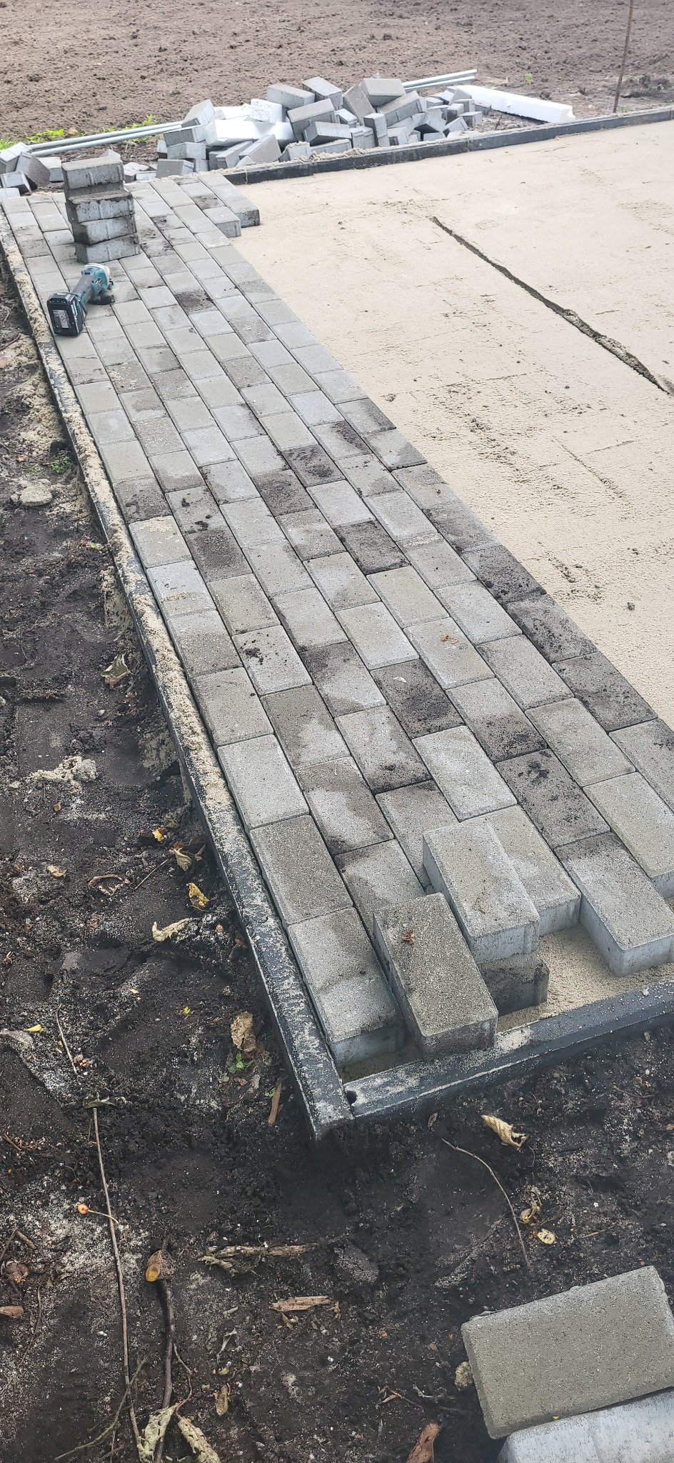 Układanie szarej kostki brukowej na podsypce piaskowej, widoczne narzędzie i zapas kostki. Krawężnik wyznacza granice. Częściowo ukończona nawierzchnia.