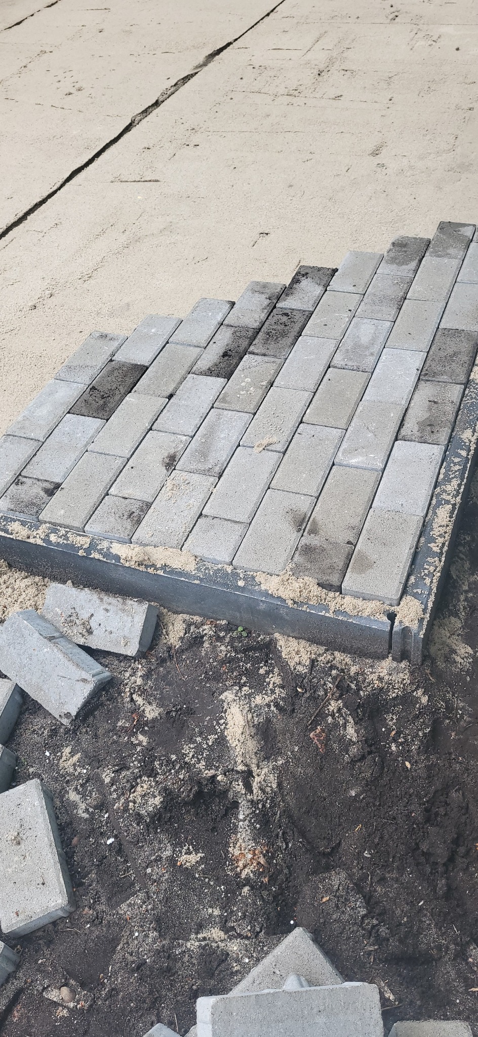 Układanie szarej kostki brukowej na podsypce piaskowej, widoczne obrzeże i zapas kostki brukowej. Etap prac ziemnych i przygotowania podłoża.