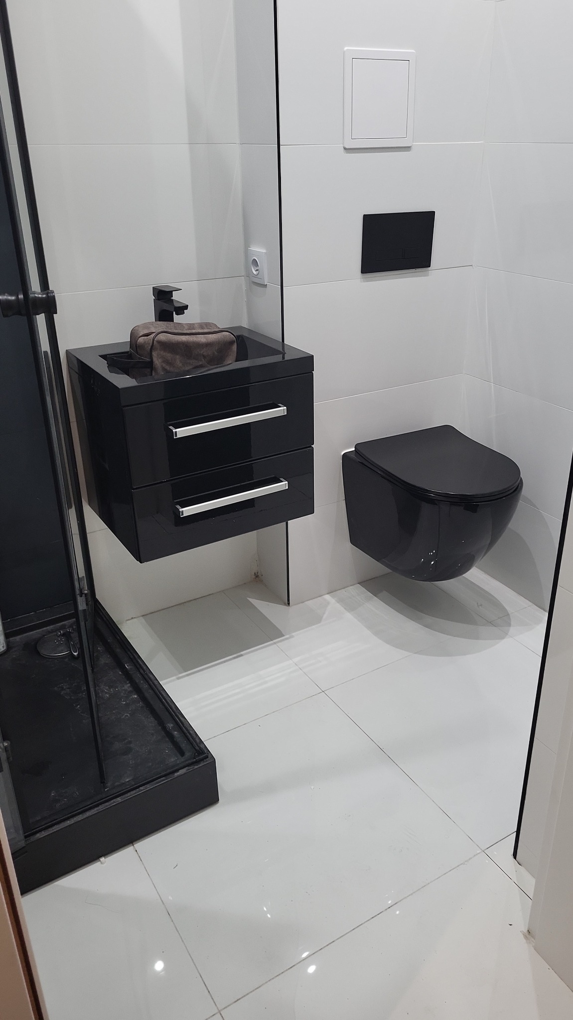 Nowoczesna łazienka z czarną armaturą: wisząca toaleta, szafka z umywalką i kabina prysznicowa. Minimalistyczny design z białymi płytkami i połyskującą podłogą.