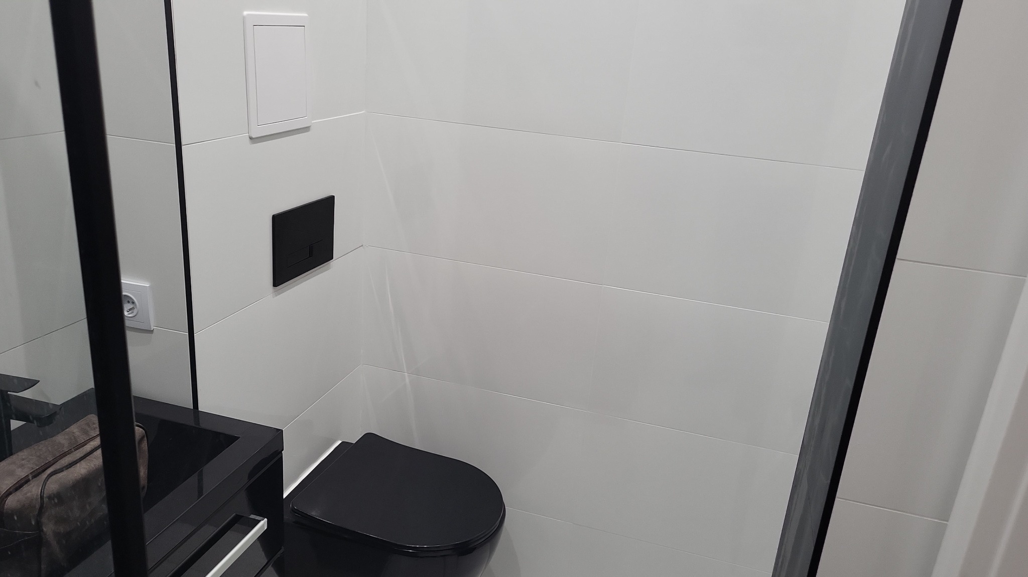 Nowoczesna toaleta z czarną armaturą i białymi płytkami. Minimalistyczny design, czarny sedes i umywalka. W tle widoczne białe płytki i czarny przycisk spłuczki.