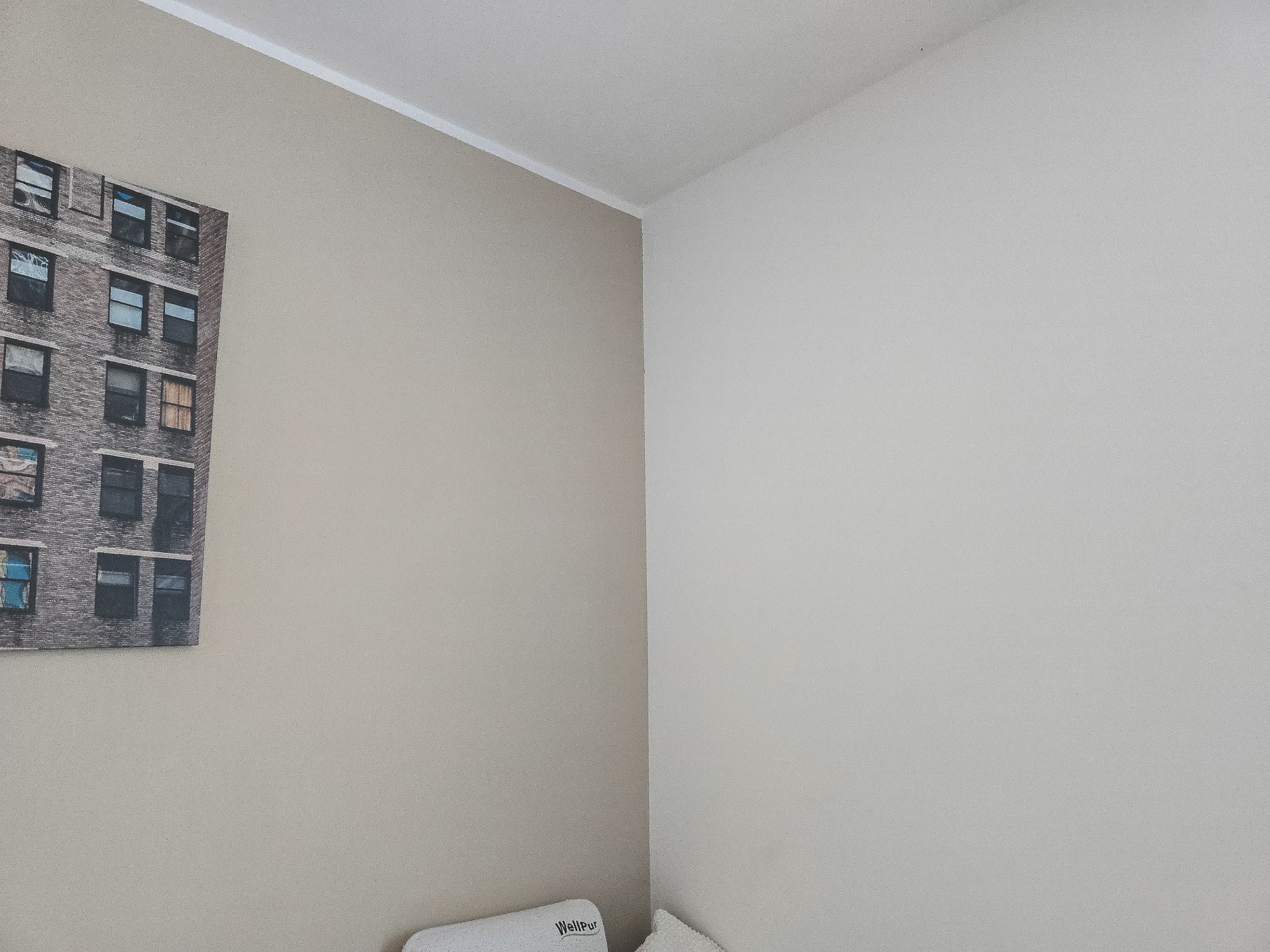 Ściana w pokoju po malowaniu, z obrazem przedstawiającym budynek. Dwie różne barwy ścian, jasna i beżowa, oddzielone narożnikiem. Widoczny fragment poduszki.