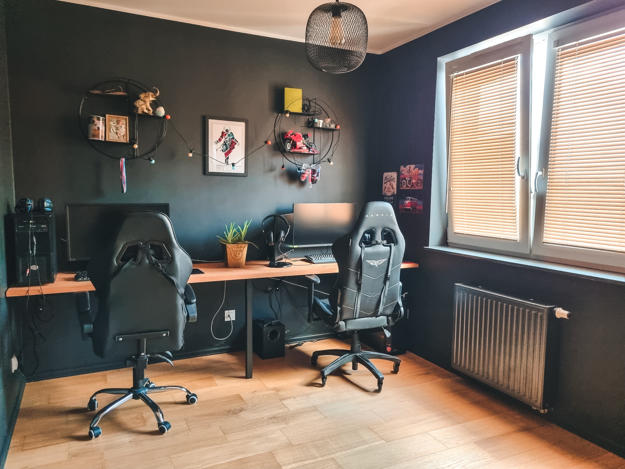 Pokój gracza po malowaniu: czarne ściany, drewniany blat biurka, dwa fotele gamingowe. Nowoczesne oświetlenie i dekoracje na ścianach.
