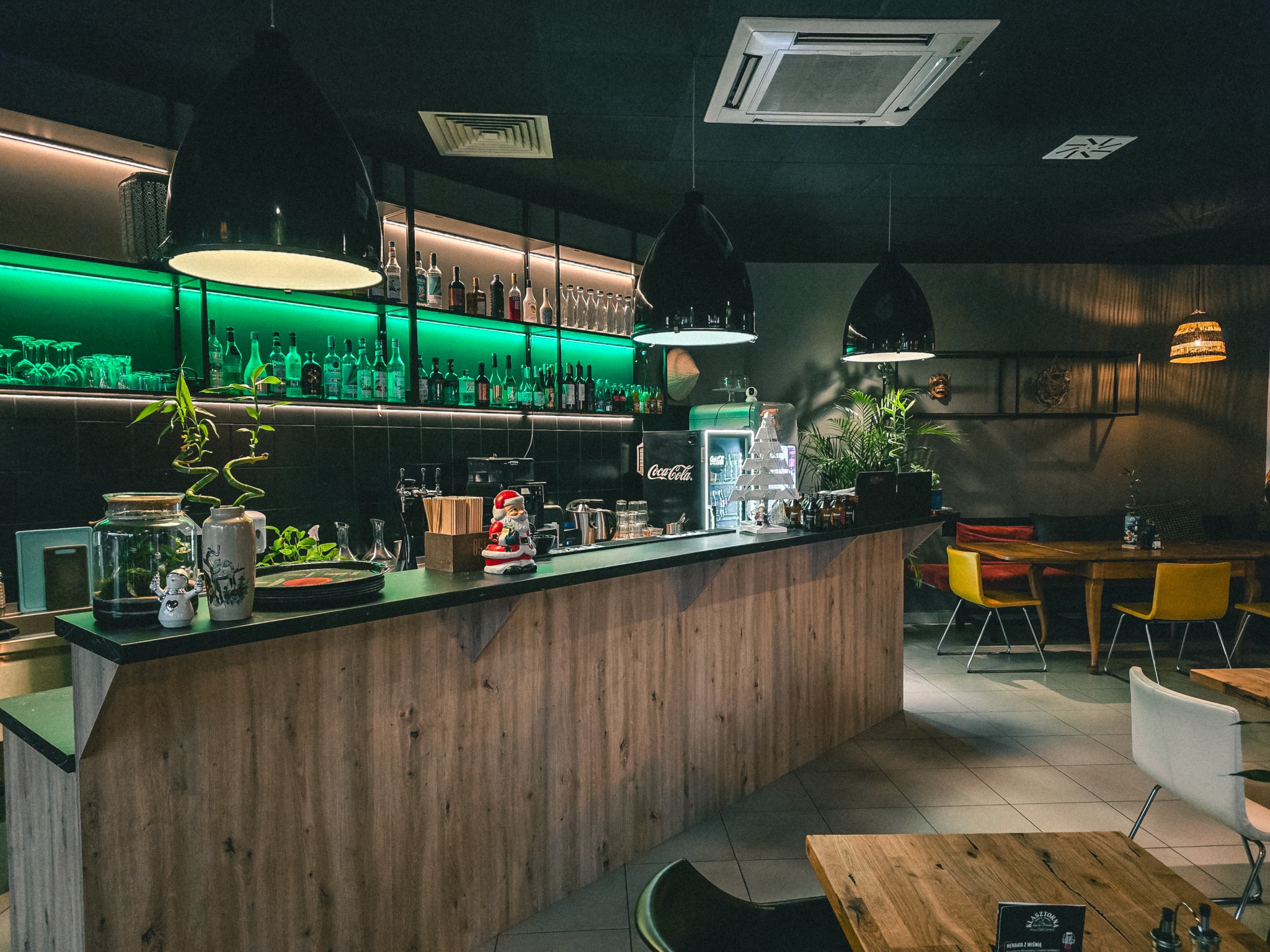 Nowoczesny bar z drewnianym blatem, podświetlanymi półkami z butelkami i oryginalnymi lampami. W tle stoliki i krzesła. Całość w ciemnej tonacji z akcentami zieleni.
