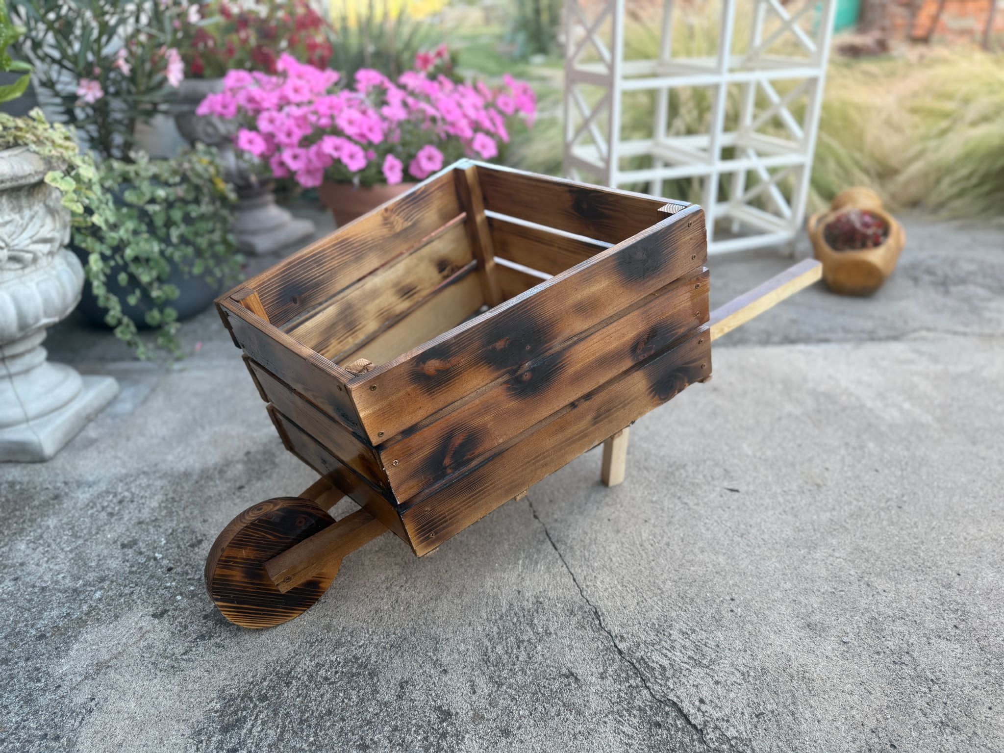 Drewniana taczka dekoracyjna z palonego drewna, ustawiona na betonowym podłożu. W tle kwiaty i elementy dekoracyjne ogrodu, tworzące rustykalny klimat.