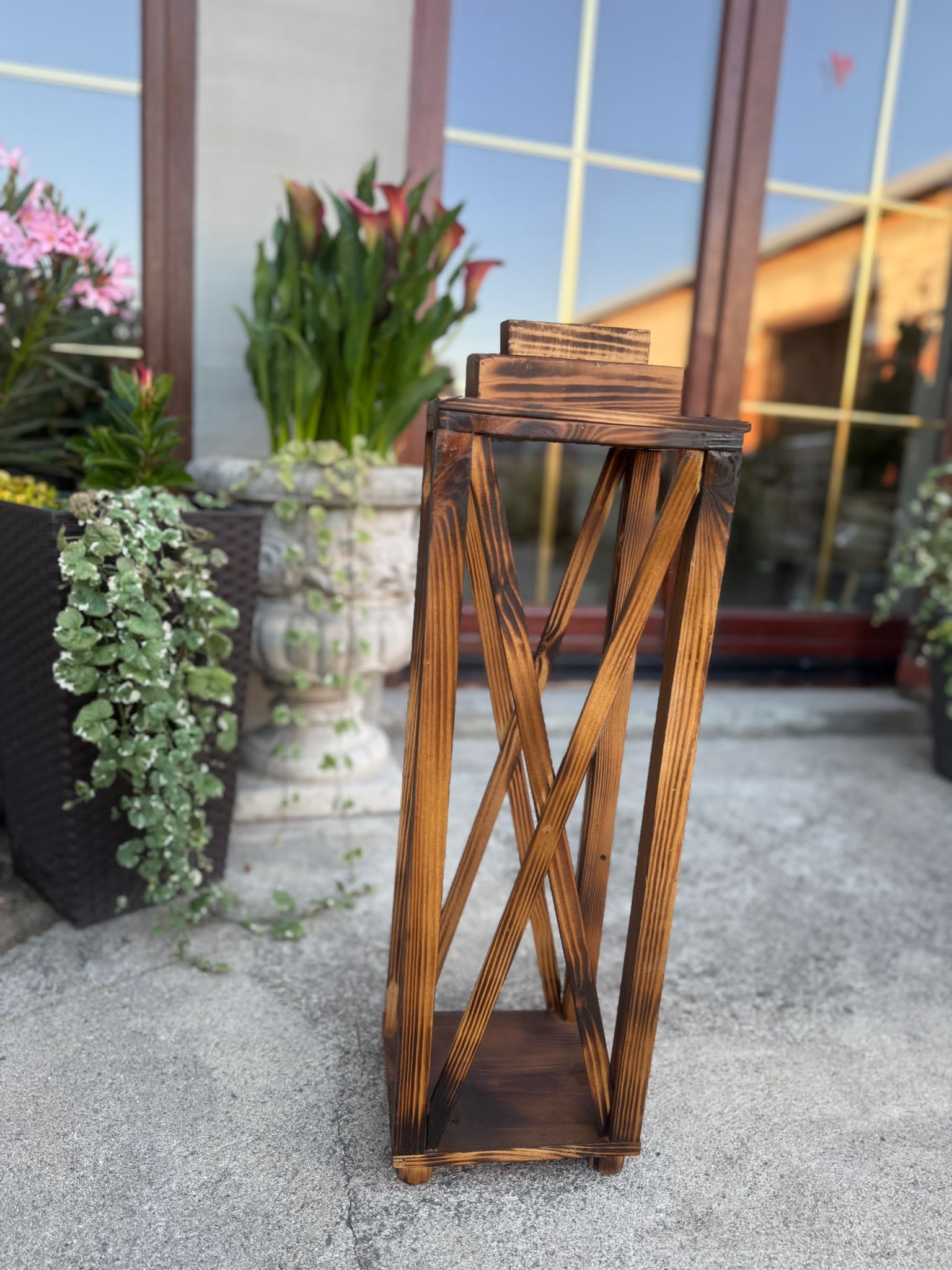 Drewniana, dekoracyjna latarenka z opalonymi brzegami, na zewnątrz, w tle rośliny w donicach i okno z refleksami. Ozdoba w stylu rustykalnym.