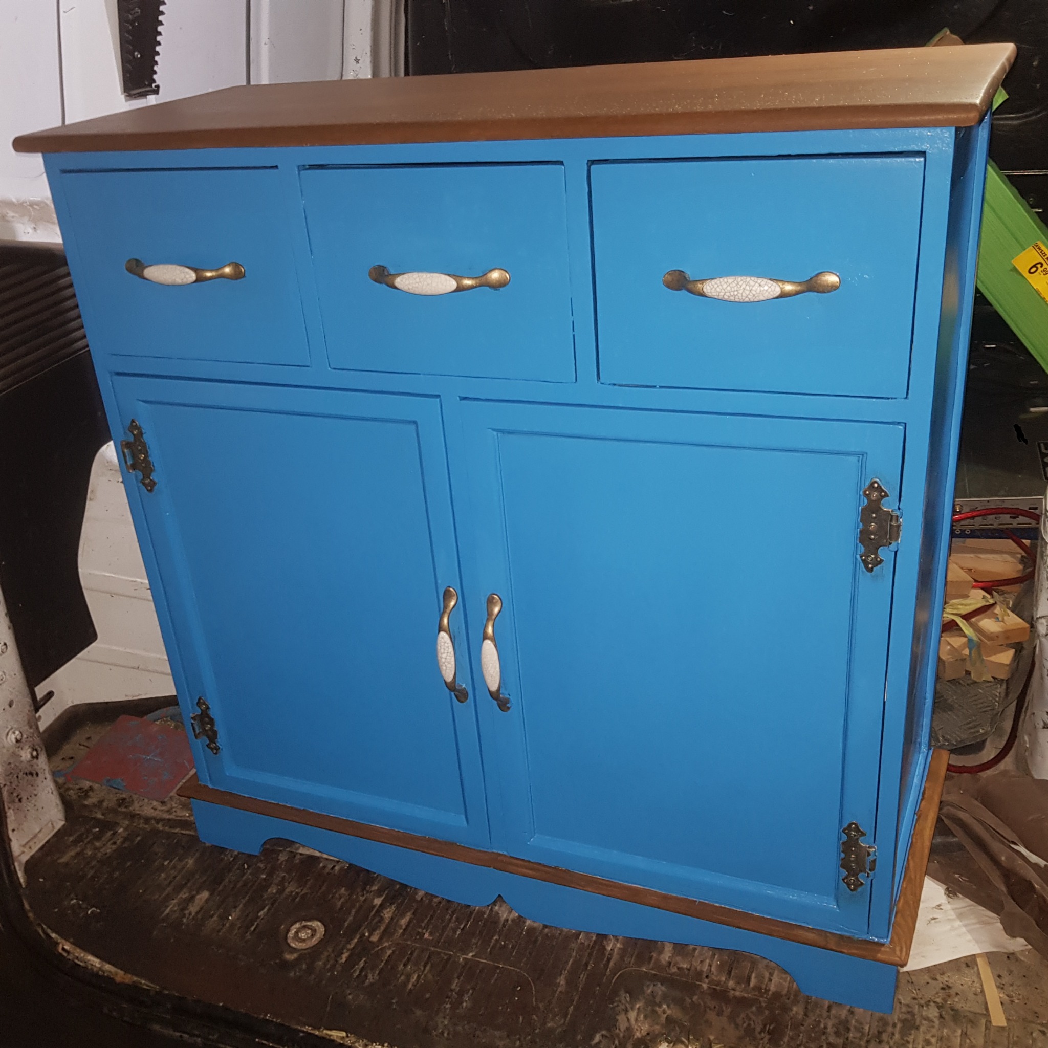 Niebieska komoda z trzema szufladami i dwojgiem drzwiczek, z ceramicznymi uchwytami i ciemnobrązowym blatem, ustawiona na podłodze w trakcie transportu.