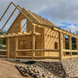 Surowy stan konstrukcji z bali z widocznym szkieletem dachu i fundamentem z kamieni polnych. Budowa w toku, na tle zieleni i błękitnego nieba.