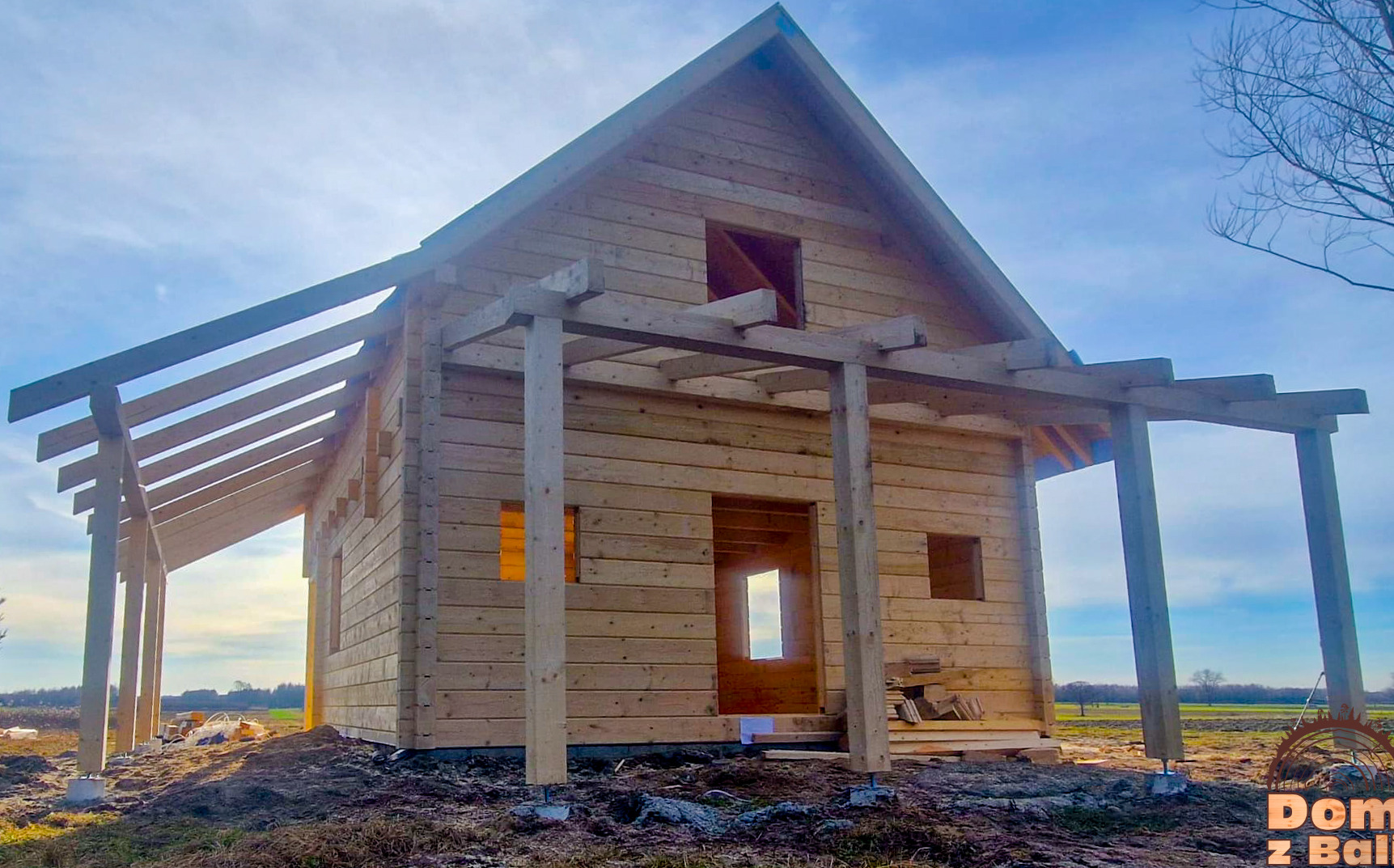 Surowy szkielet drewnianego domu z bali w trakcie budowy, z widoczną konstrukcją dachu i zadaszeniem werandy, na tle pola i błękitnego nieba.
