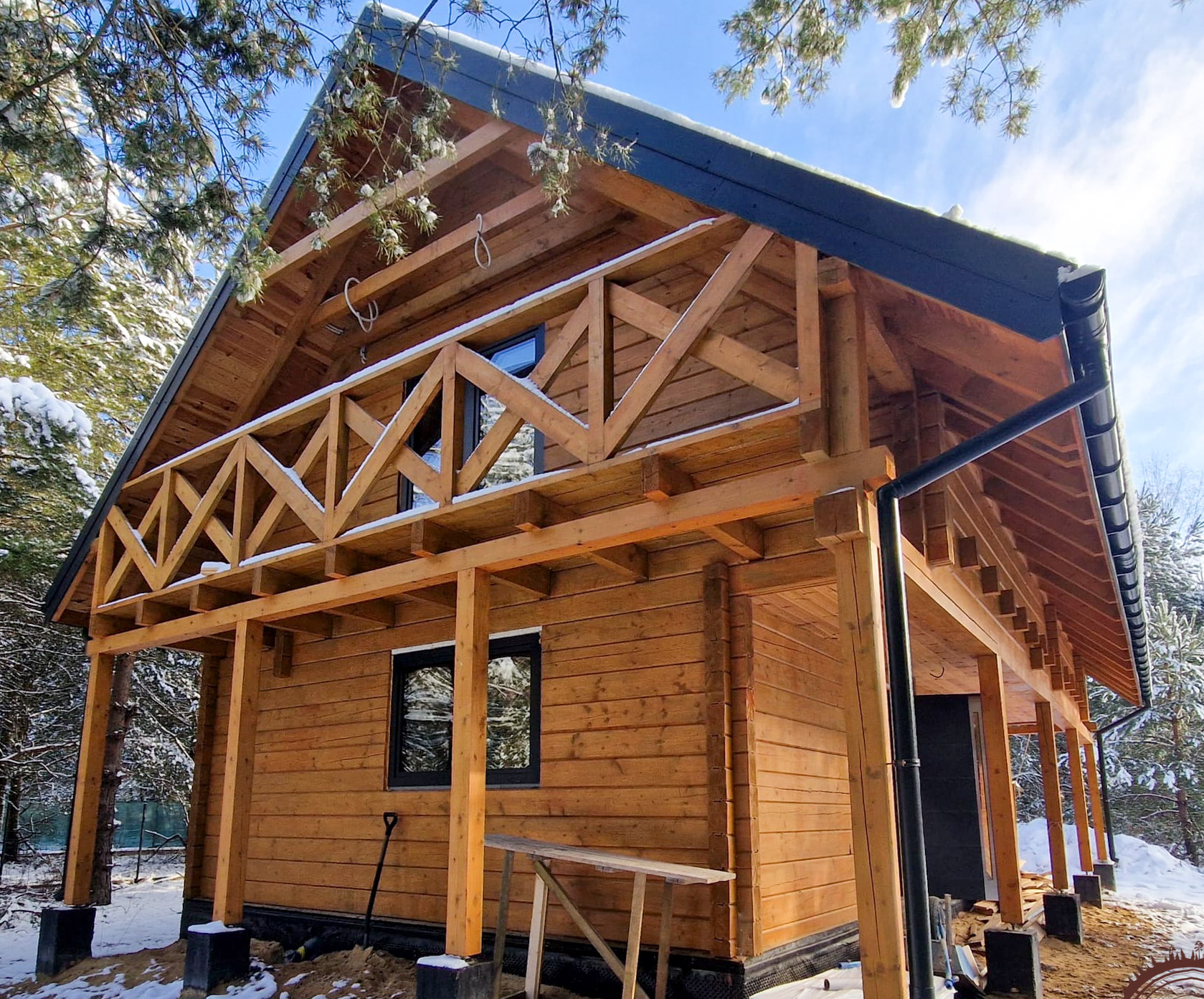 Surowy dom z bali w zimowej scenerii, z widoczną konstrukcją balkonu i elementami wykończeniowymi, takimi jak rynny. Widoczny śnieg na drzewach i dachu.