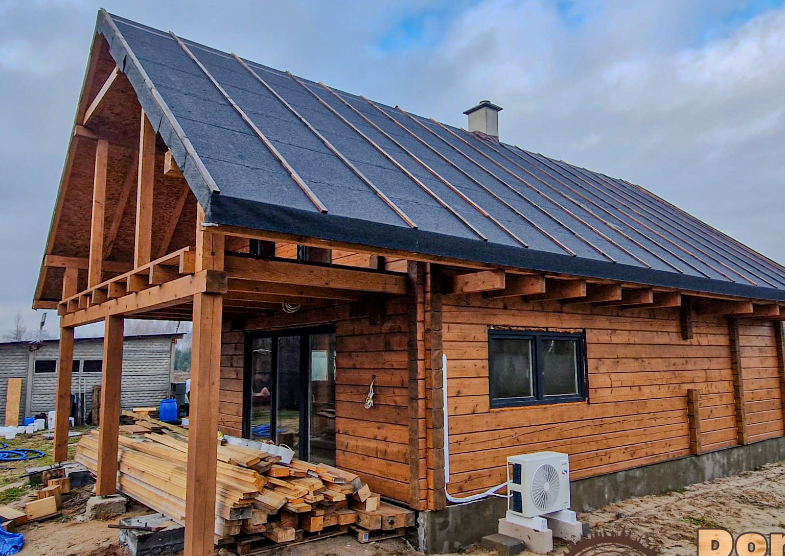 Budowa domu z bali w trakcie: drewniana konstrukcja, dach pokryty papą, klimatyzator na zewnątrz. Widoczne deski i elementy konstrukcyjne.