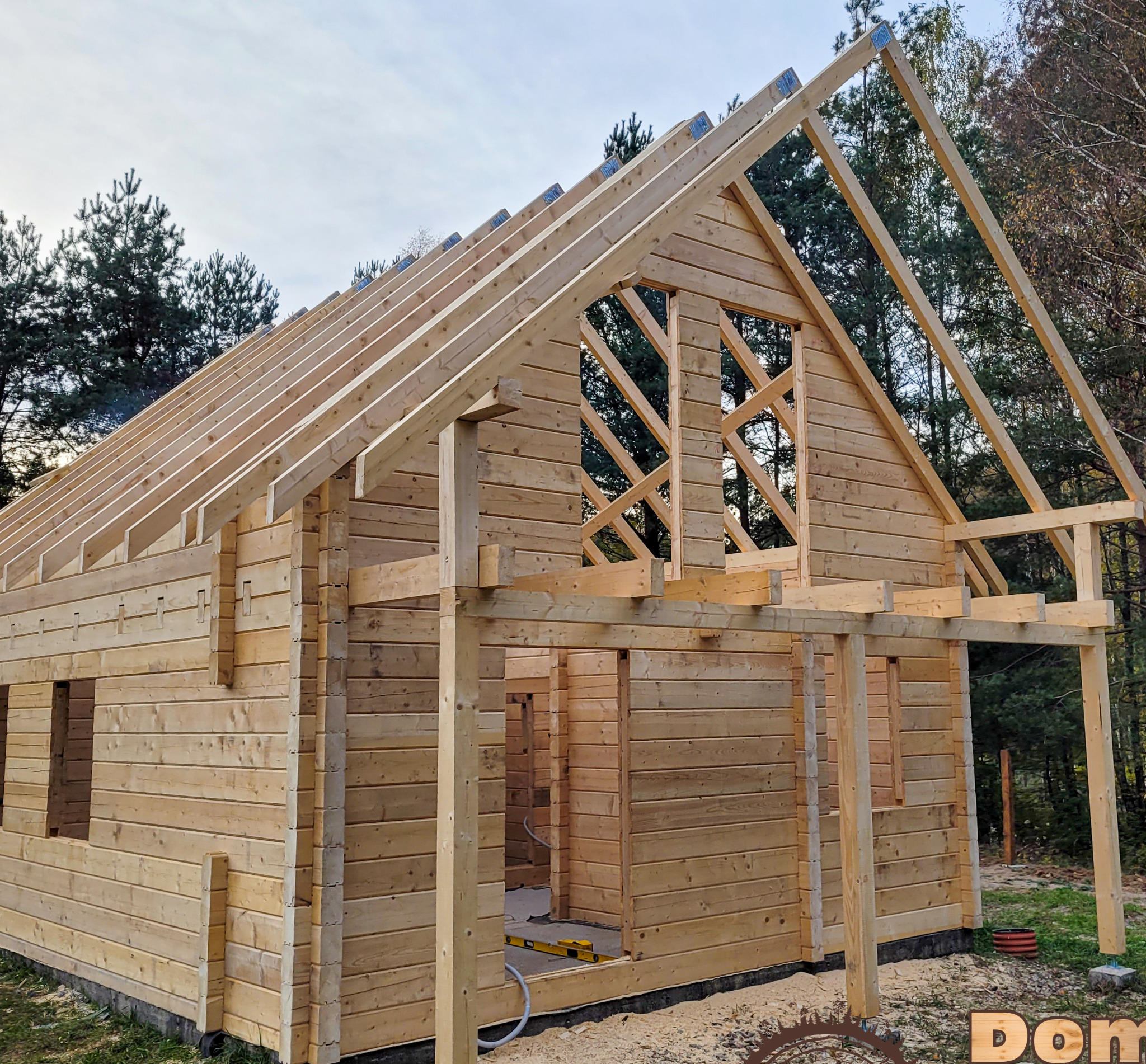 Surowy szkielet domu z bali w trakcie budowy, widoczne drewniane ściany, konstrukcja dachu i zarys przyszłego wejścia na tle drzew i pochmurnego nieba.