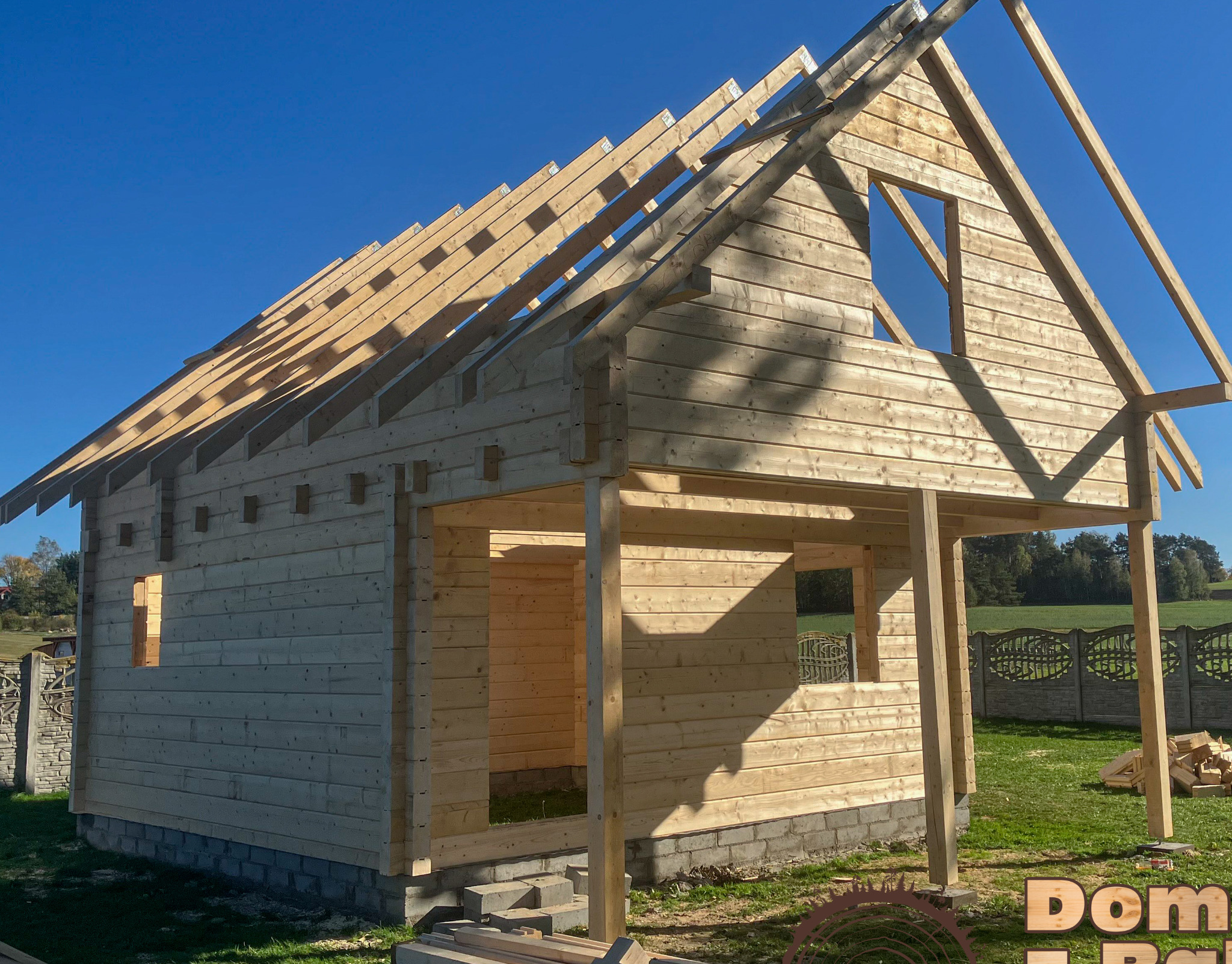 Surowy dom z bali w trakcie budowy, widoczne ściany z bali, krokwie dachowe i otwory okienne na tle błękitnego nieba i zielonej łąki.