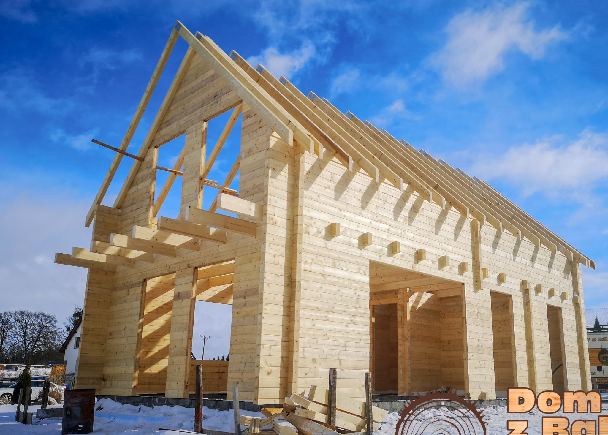 Surowy szkielet domu z bali w zimowej scenerii, ukazujący konstrukcję ścian i dachu na tle błękitnego nieba z chmurami. Widoczne detale łączeń drewnianych elementów.