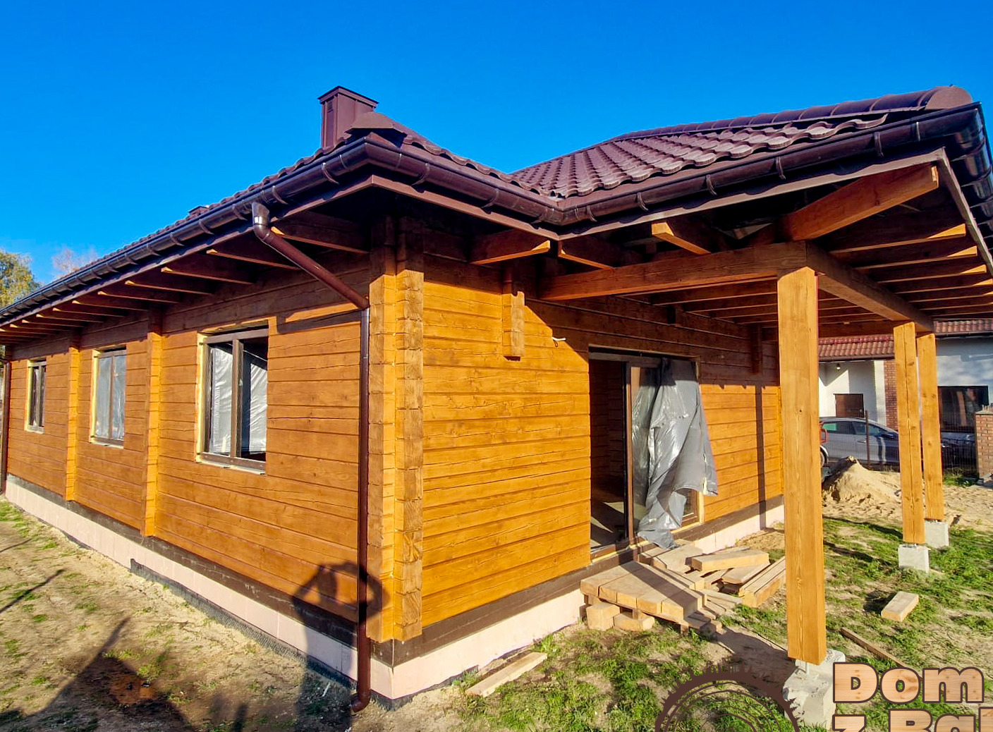 Surowy, drewniany dom z bali z brązowym dachem w Izabelinie w słoneczny dzień. Widoczny fragment tarasu i okna zabezpieczone folią, co sugeruje etap wykończeniowy.