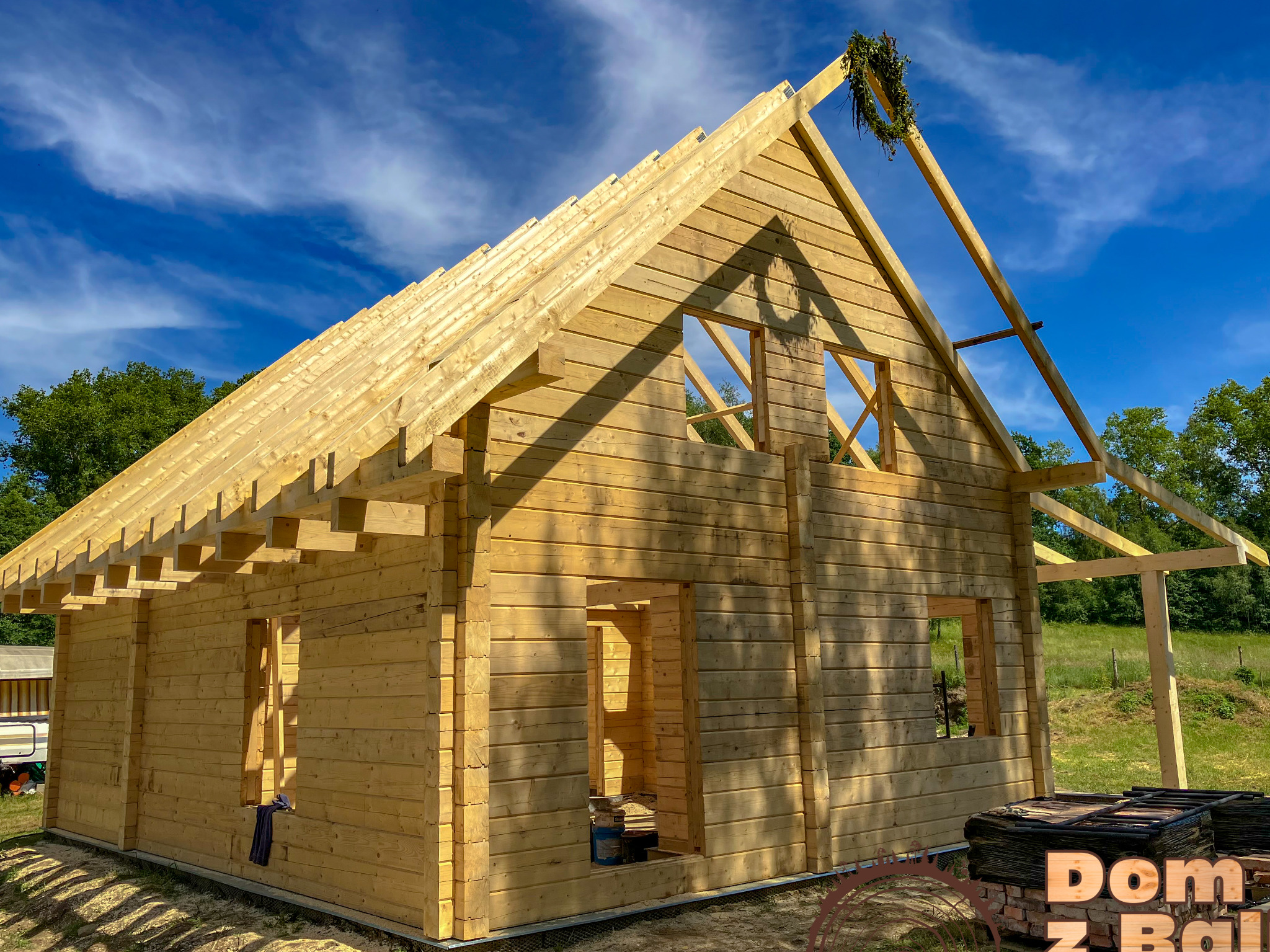 Surowy stan domu z bali, widoczne belki stropowe i wieniec na dachu. Błękitne niebo w tle, otoczenie zielone. Częściowo ukończona konstrukcja, brak okien i drzwi.