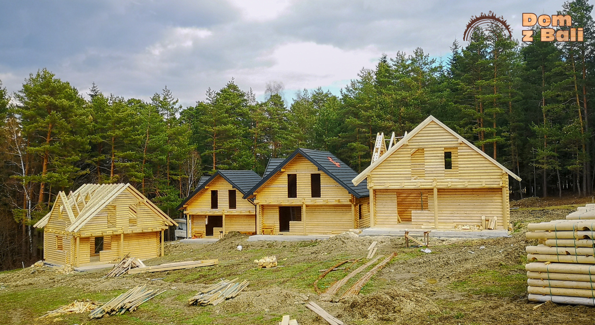 Trzy domy w różnym stanie budowy, wykonane z jasnych bali, na tle lasu iglastego. Jeden dom z dachem w trakcie budowy, drugi z czarną dachówką.