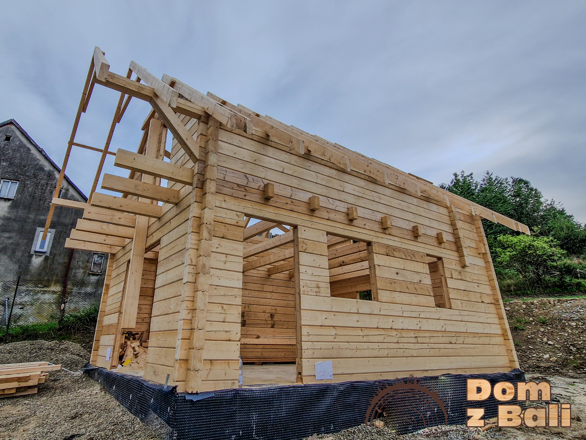 Surowy szkielet drewnianego domu w budowie, z widocznymi belkami stropowymi i ścianami z bali, na tle pochmurnego nieba i sąsiedniego budynku.