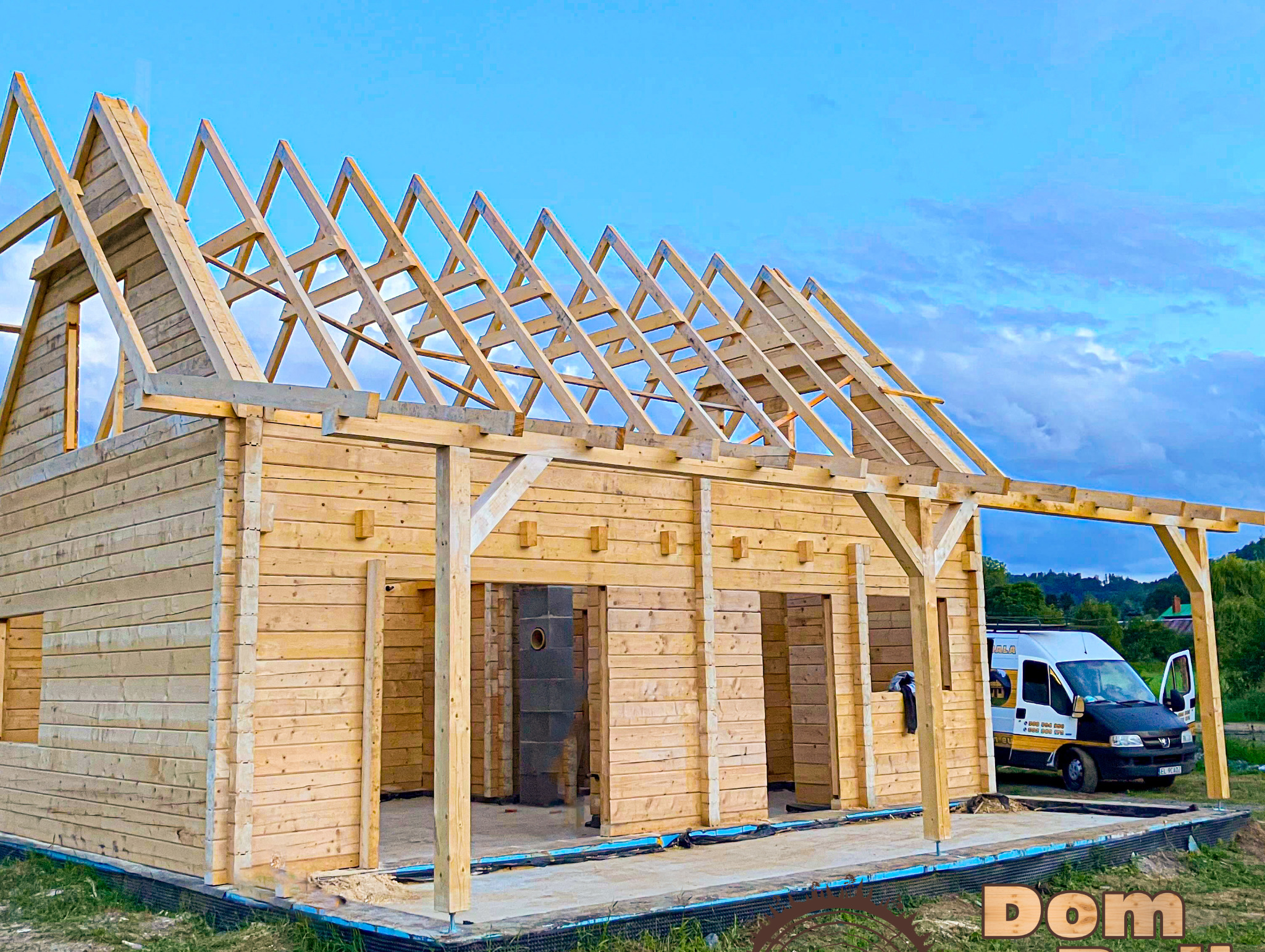 Drewniany dom w trakcie budowy, widoczne ściany z bali i konstrukcja dachu, na zewnątrz zaparkowany bus firmy budowlanej.