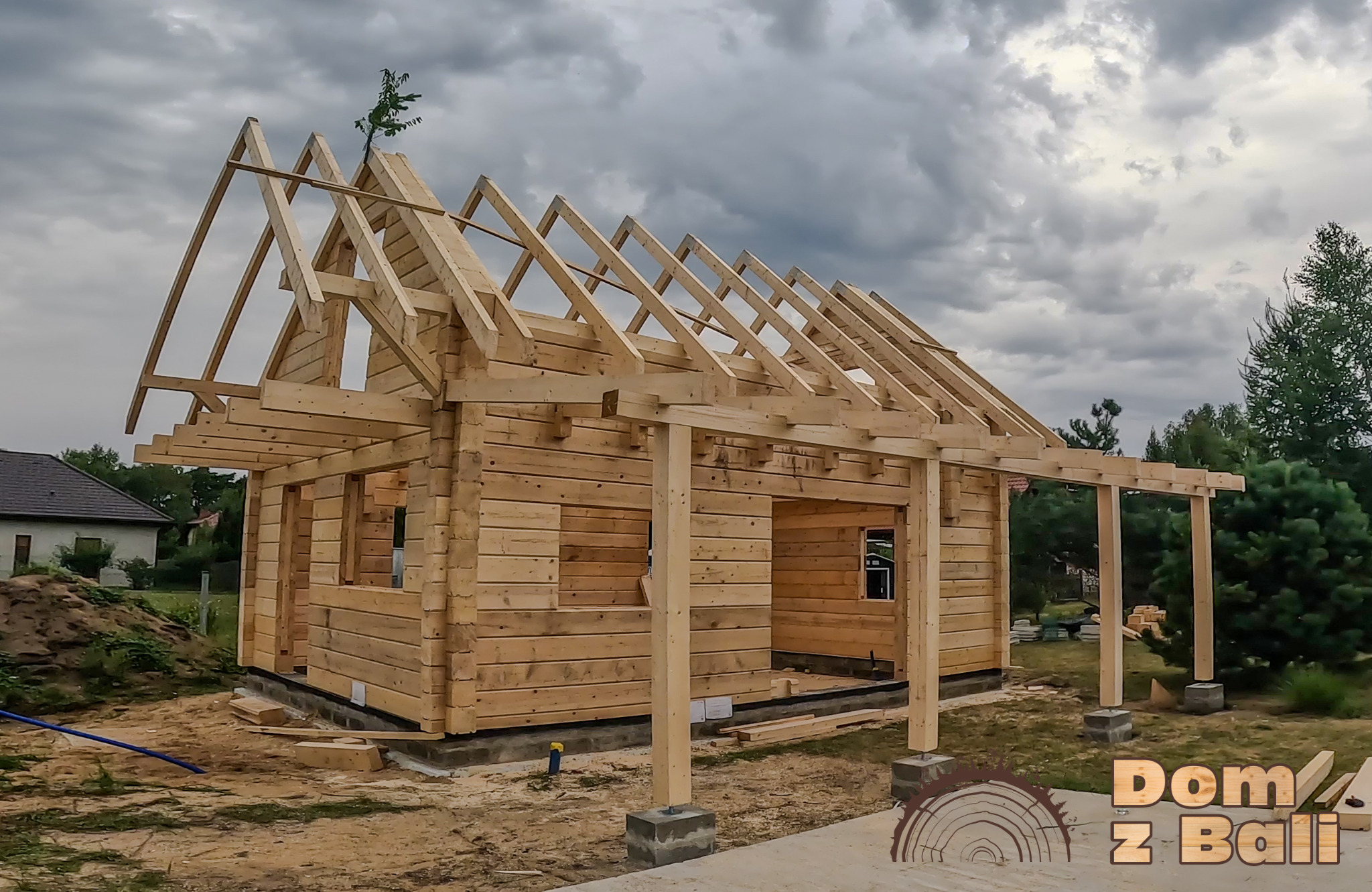 Surowy dom z bali w trakcie budowy, widoczne drewniane ściany, krokwie dachu i słupy werandy. W tle pochmurne niebo i dom w oddali. Na szczycie dachu małe drzewko.