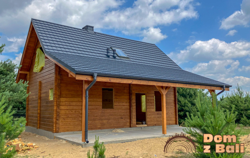 Surowy stan budowy domu z bali z ciemną dachówką i zadaszonym tarasem, widok z zewnątrz w słoneczny dzień. Część okien zakryta folią.