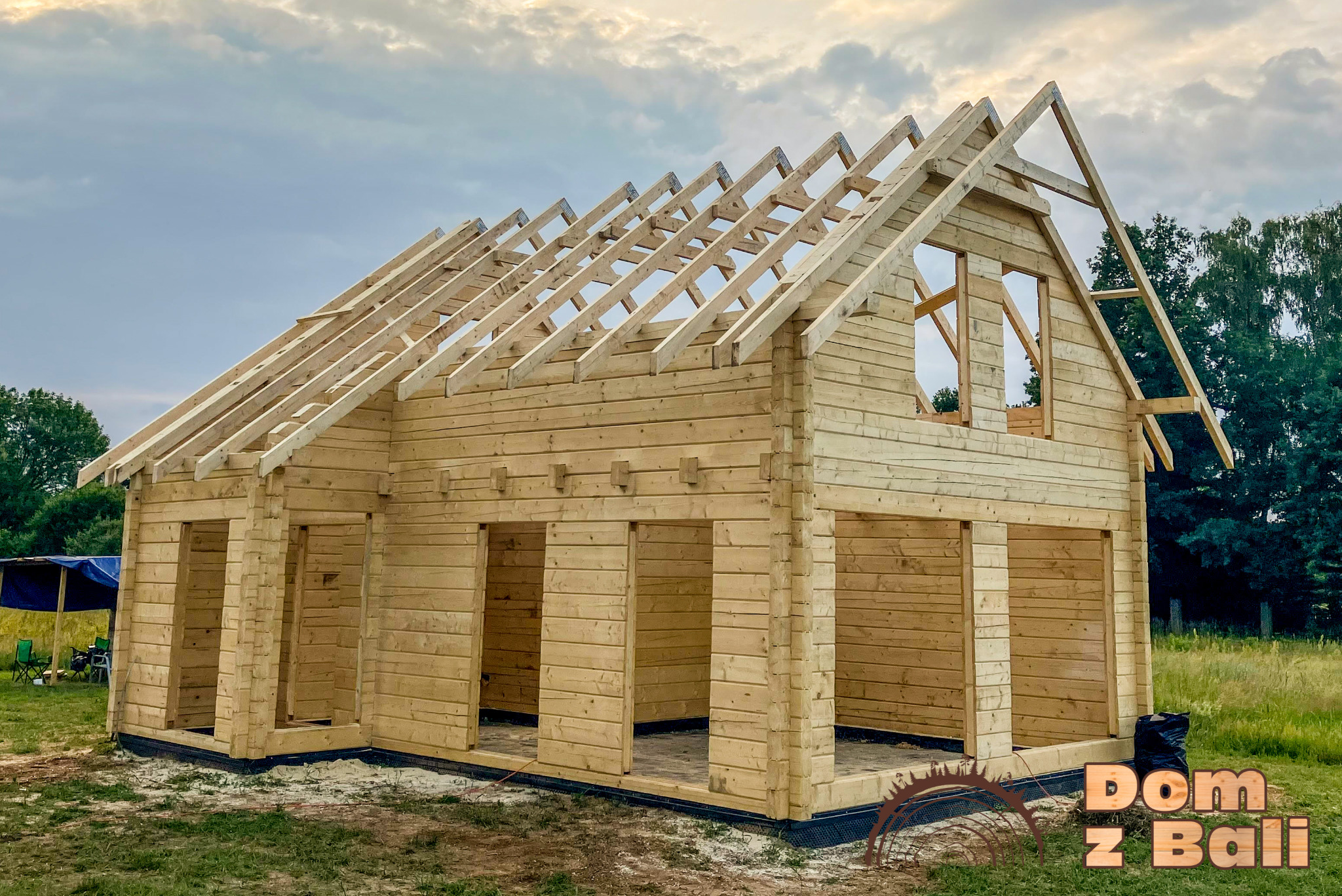 Surowy szkielet domu z bali w trakcie budowy, widoczne drewniane ściany i konstrukcja dachu na tle nieba i zieleni. Na dole widoczne logo firmy.