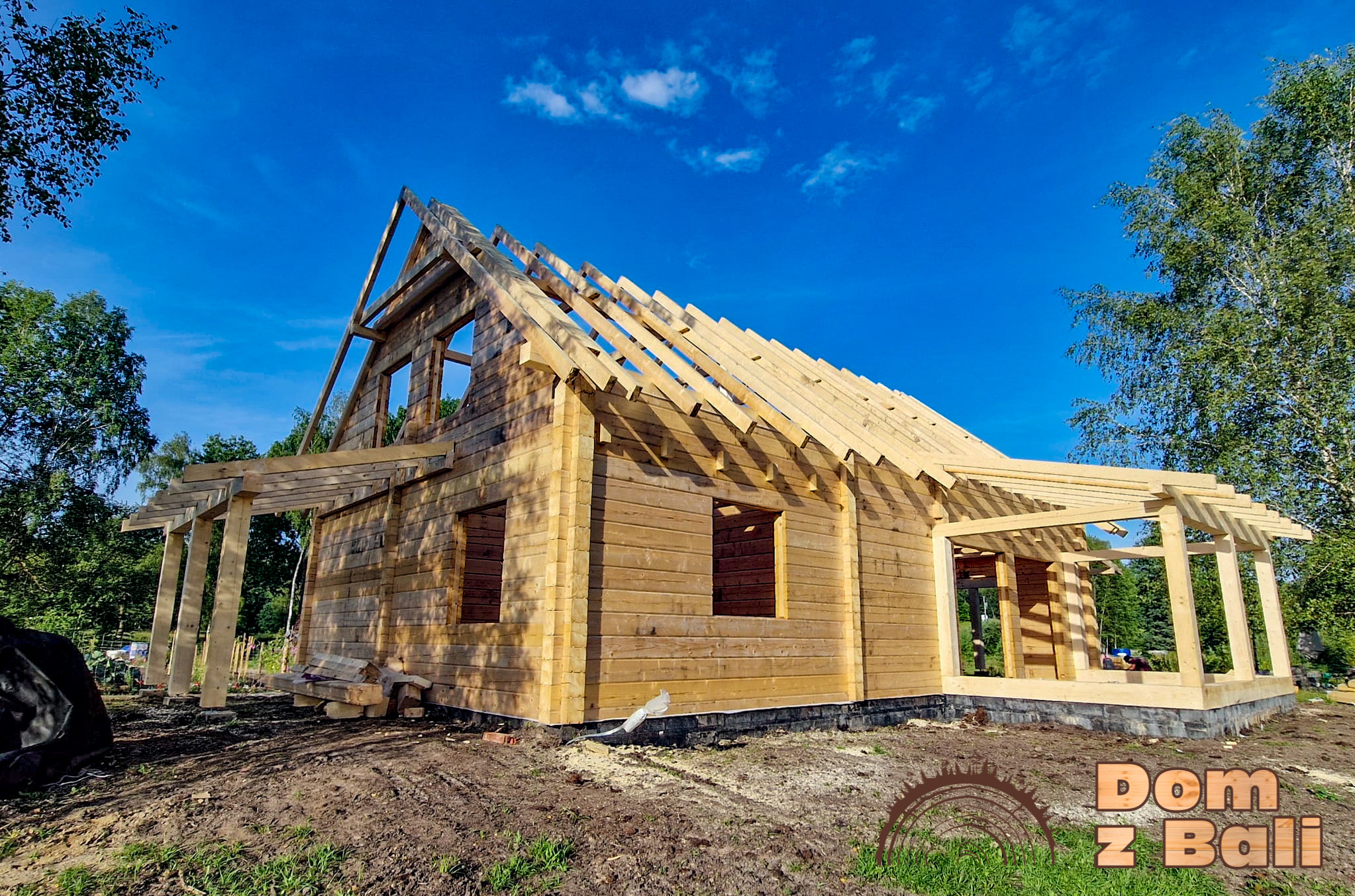 Surowy stan budowy domu z bali, widoczne drewniane ściany, konstrukcja dachu i tarasu na tle błękitnego nieba i zieleni drzew. Dom w trakcie wznoszenia.