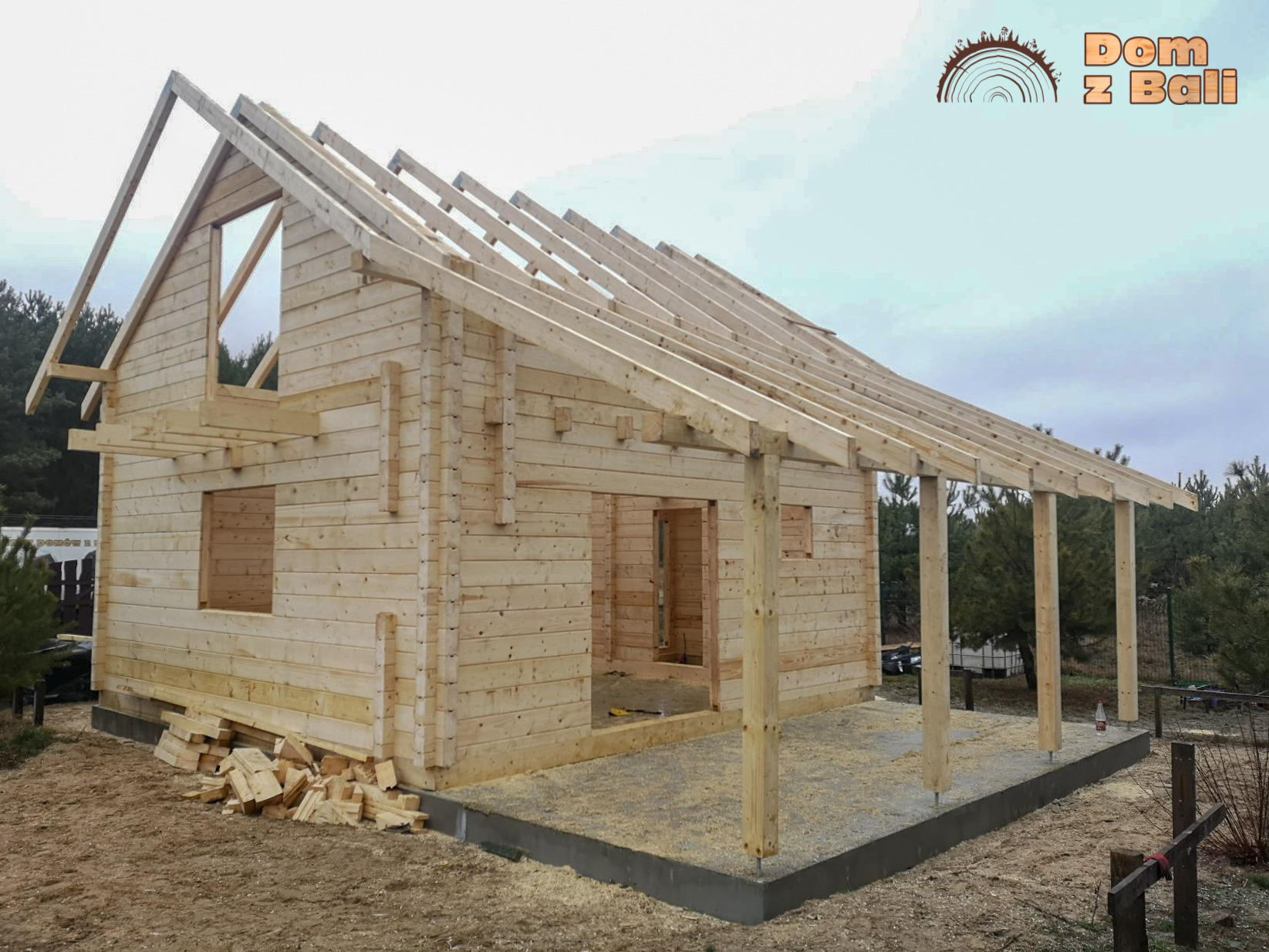 Surowy stan budowy domu z bali: drewniana konstrukcja szkieletowa z widocznym zadaszeniem werandy, fundamentem i stertą odpadów drewnianych. W tle las.