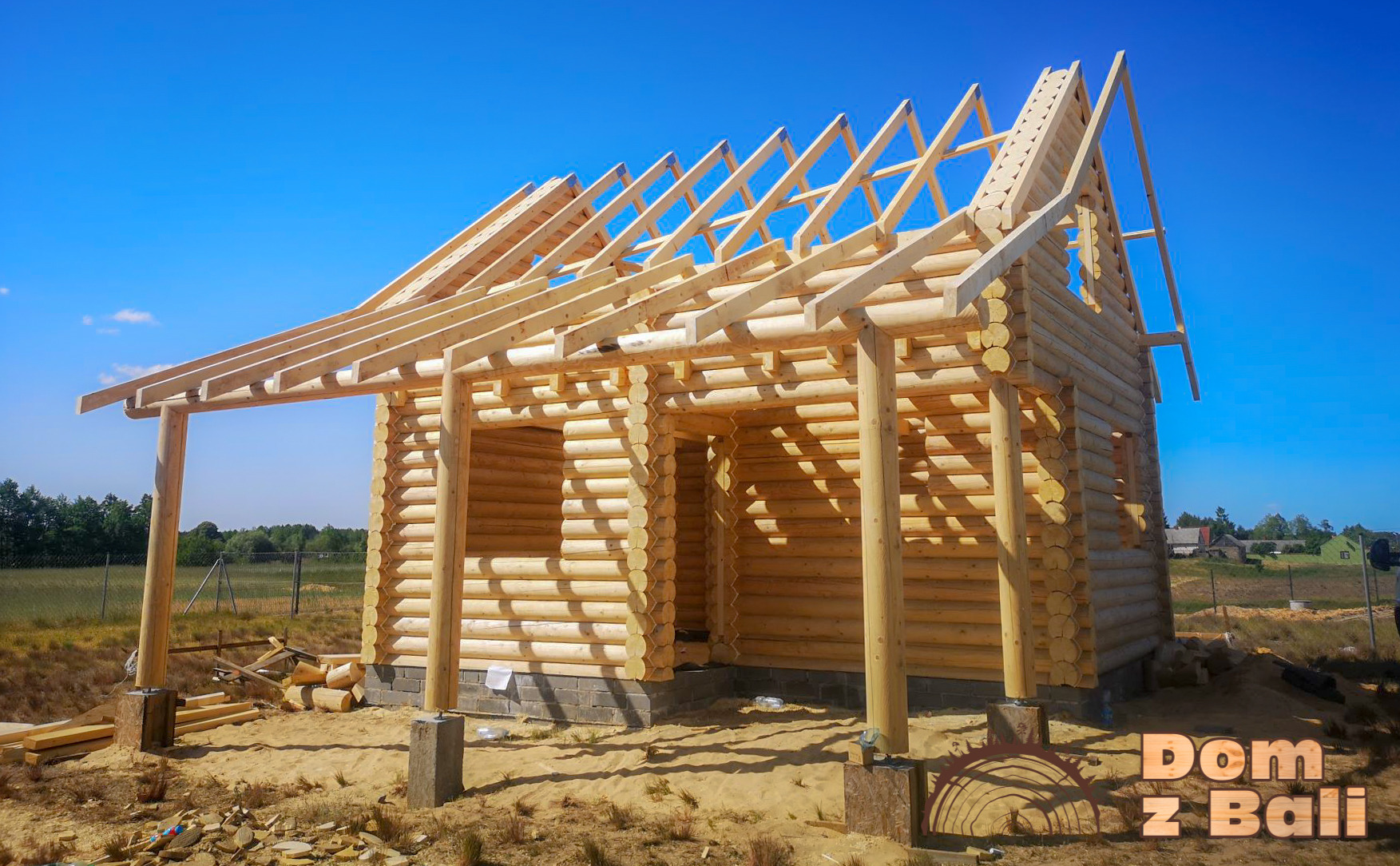 Surowy szkielet domu z bali w budowie, widoczne drewniane ściany, krokwie dachowe na tle błękitnego nieba, fundamenty z bloczków betonowych.