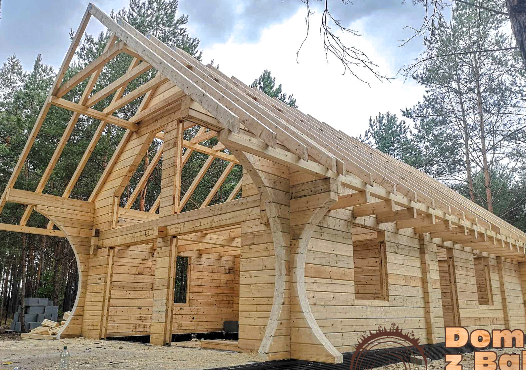 Konstrukcja domu z bali w trakcie budowy, widoczne drewniane ściany, więźba dachowa i elementy wykończeniowe. W tle drzewa i pochmurne niebo.