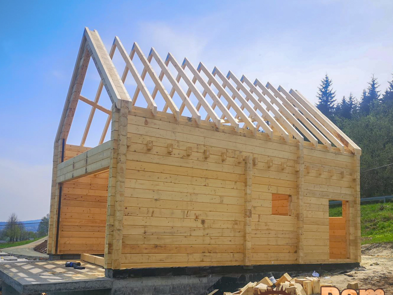 Konstrukcja domu z bali na fundamencie, widoczne krokwie dachowe, tło zielone wzgórza i błękitne niebo. Dom w stanie surowym otwartym.