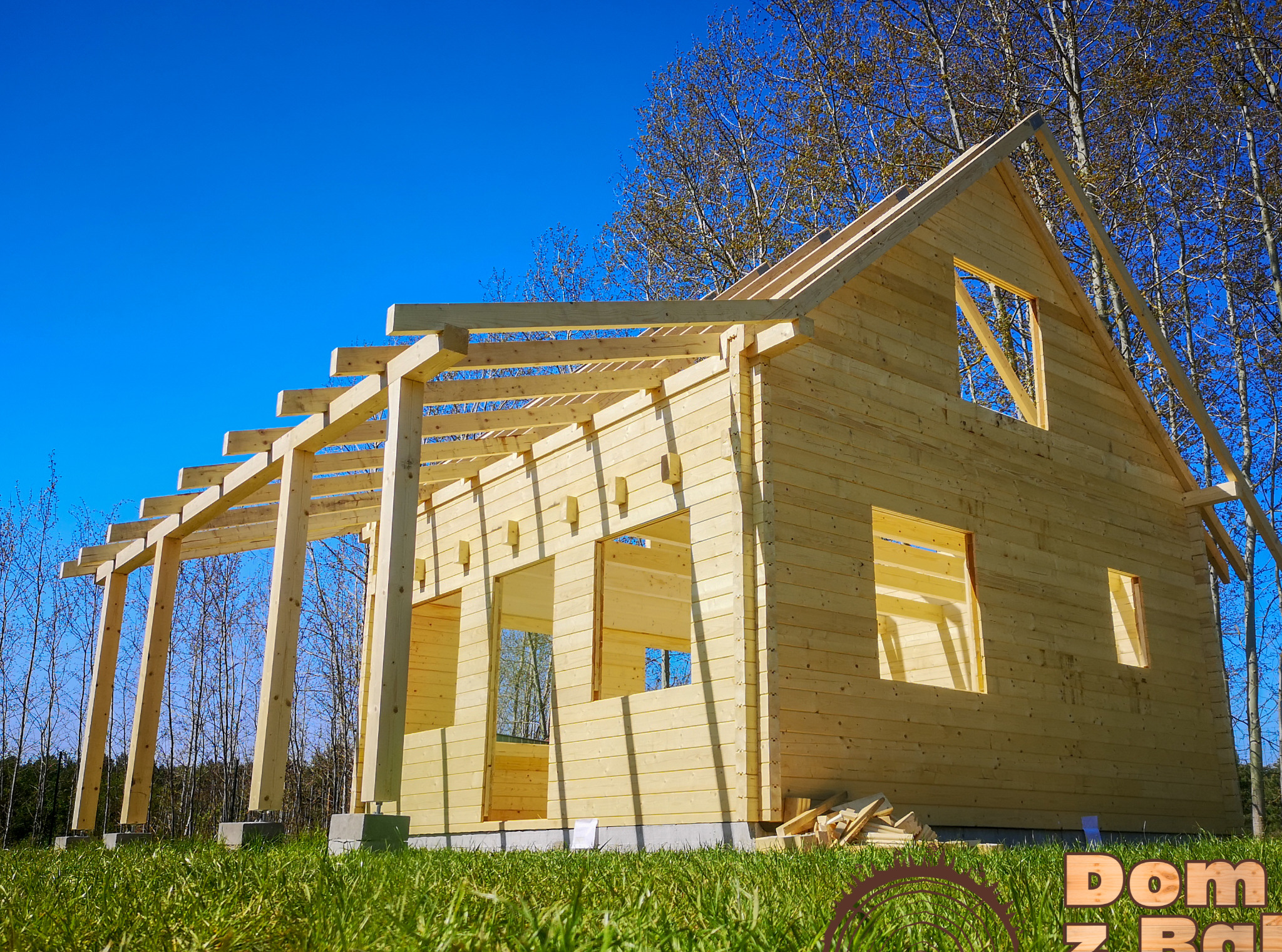 Surowy stan drewnianego domu z bali w trakcie budowy, z widoczną konstrukcją dachu i ścian na tle błękitnego nieba i drzew.