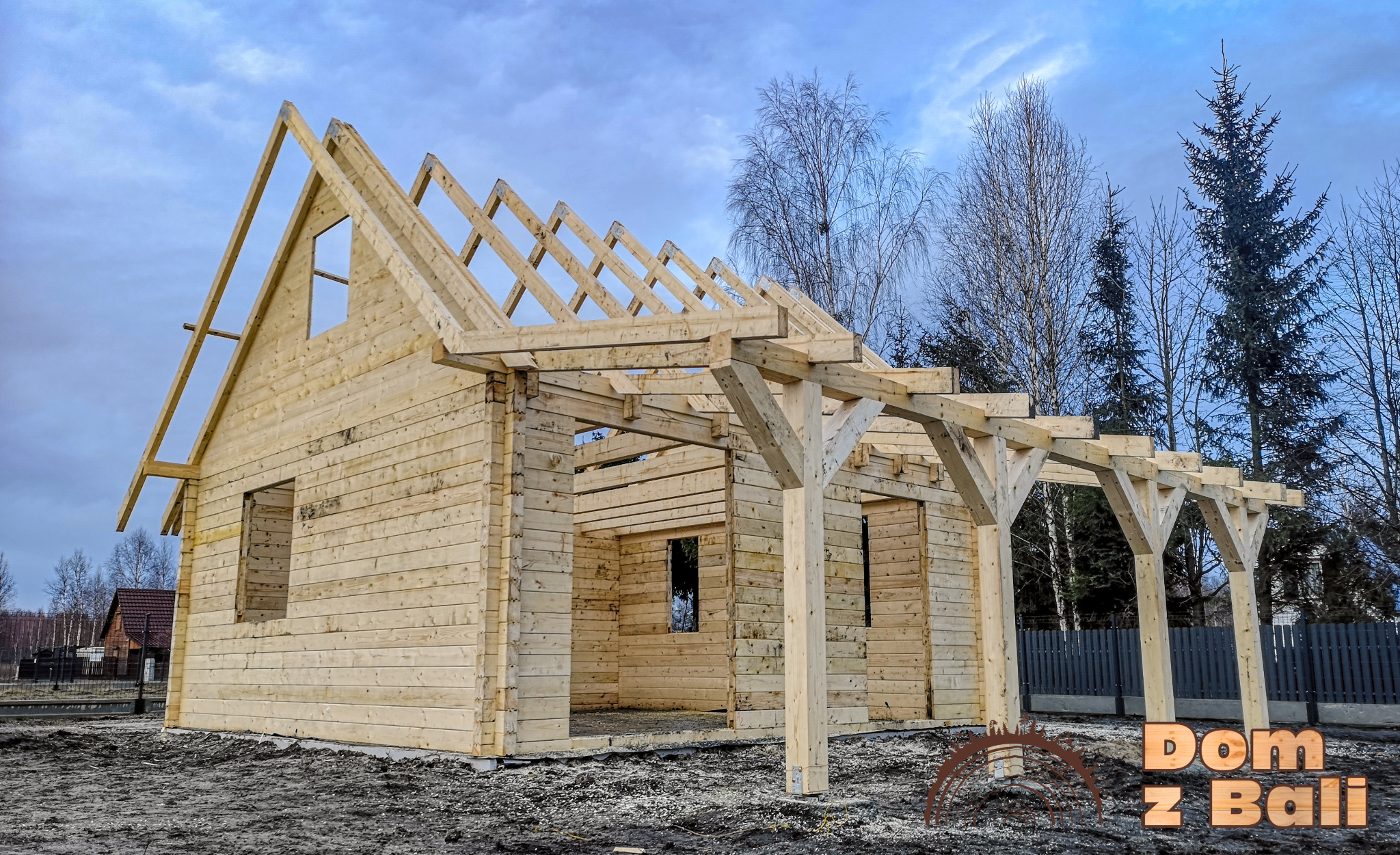 Konstrukcja surowego, jasnego domu z bali w trakcie budowy, z widocznym szkieletem dachu na tle pochmurnego nieba i drzew. Widoczne detale łączeń belek.