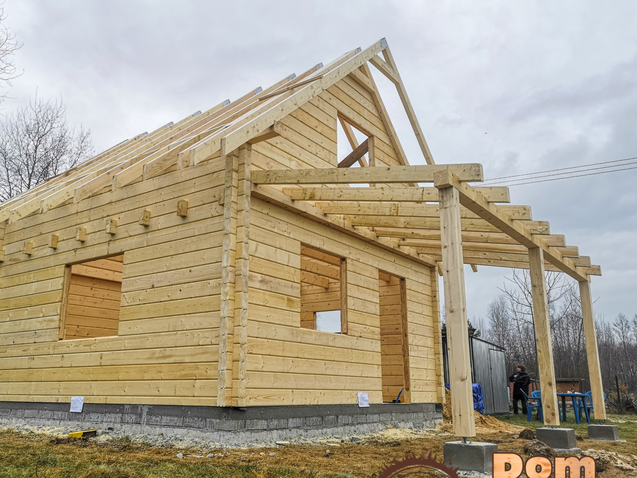 Konstrukcja domu z bali w trakcie budowy, widoczne ściany, więźba dachowa, słupy podtrzymujące zadaszenie tarasu, na zewnątrz osoba i meble ogrodowe.