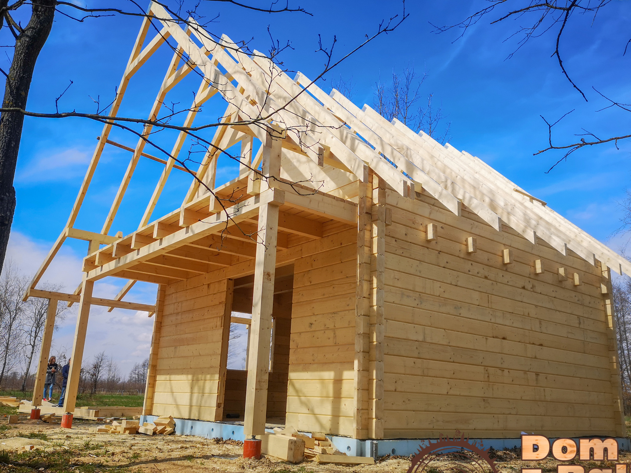 Konstrukcja domu z bali w trakcie budowy, widoczne drewniane ściany i szkielet dachu na tle błękitnego nieba z gałęziami drzew na pierwszym planie.