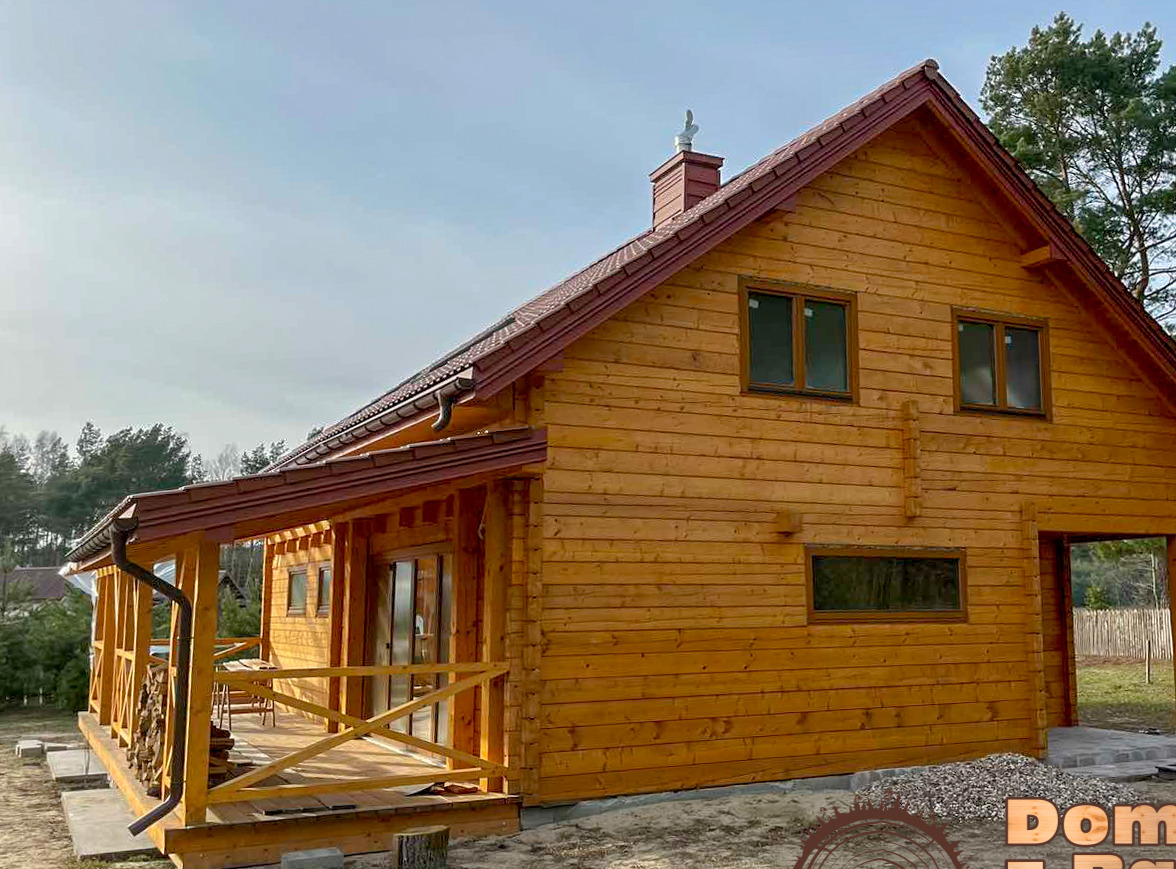 Żółty dom z bali z brązowym dachem w stylu skandynawskim, z zadaszonym tarasem. Widoczny komin z ozdobną figurką i otoczenie leśne.