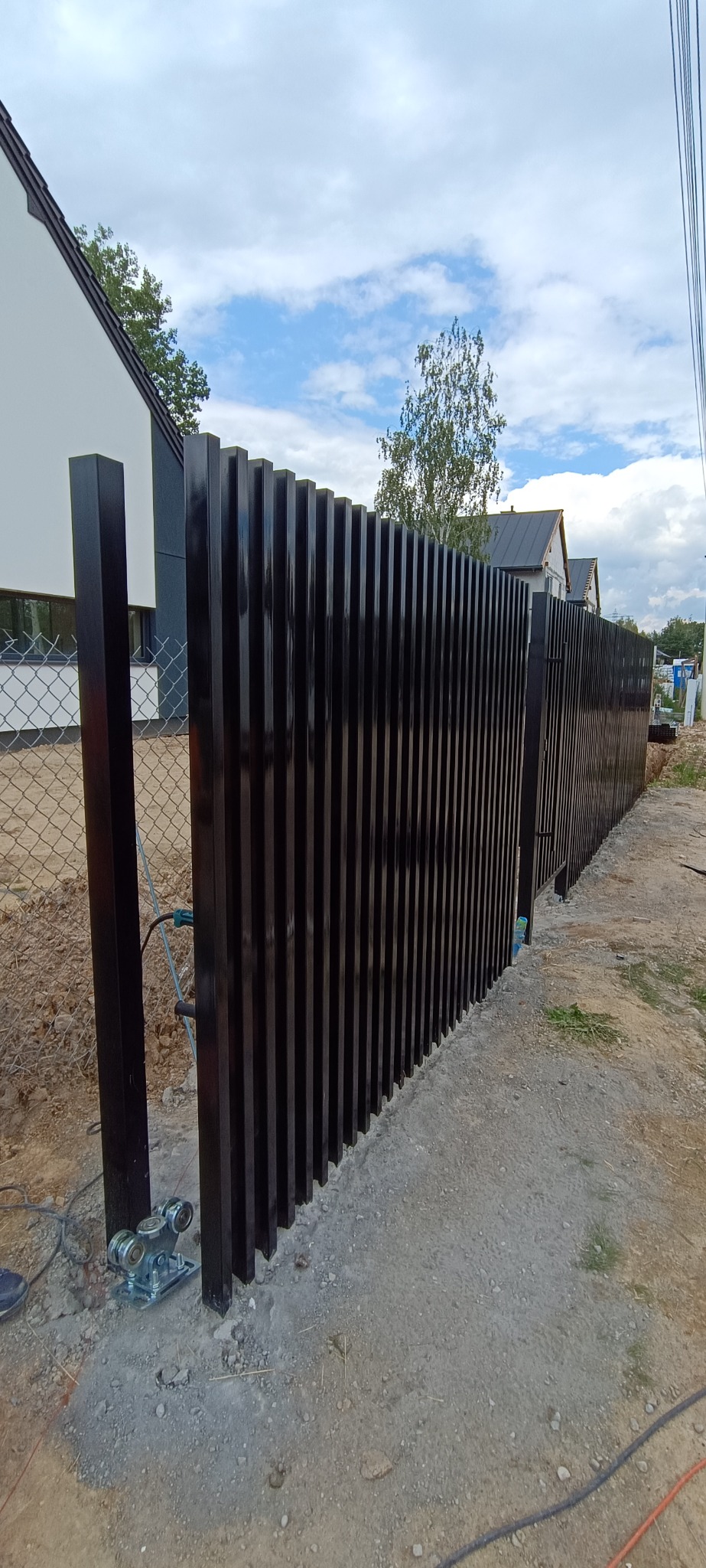 Nowoczesna, czarna brama przesuwna z pionowych profili, zamontowana na posesji. Widoczne rolki i elementy automatyki. W tle dom i błękitne niebo.