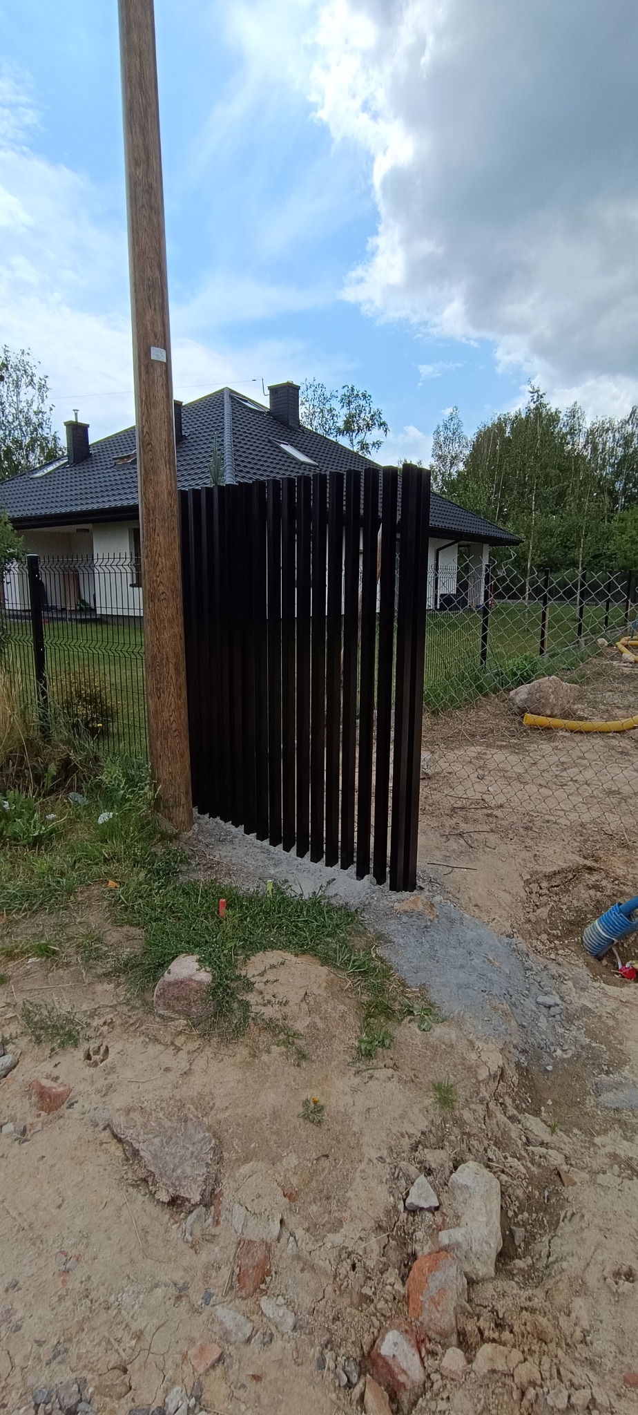 Fragment metalowego ogrodzenia panelowego w kolorze grafitowym, zamontowanego przy betonowym słupku. W tle dom i drzewa.