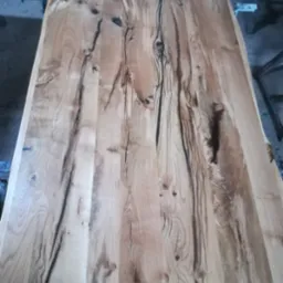 Surowy blat drewniany z wyraźnymi słojami i czarnymi pęknięciami, leżący na metalowych podporach w warsztacie.