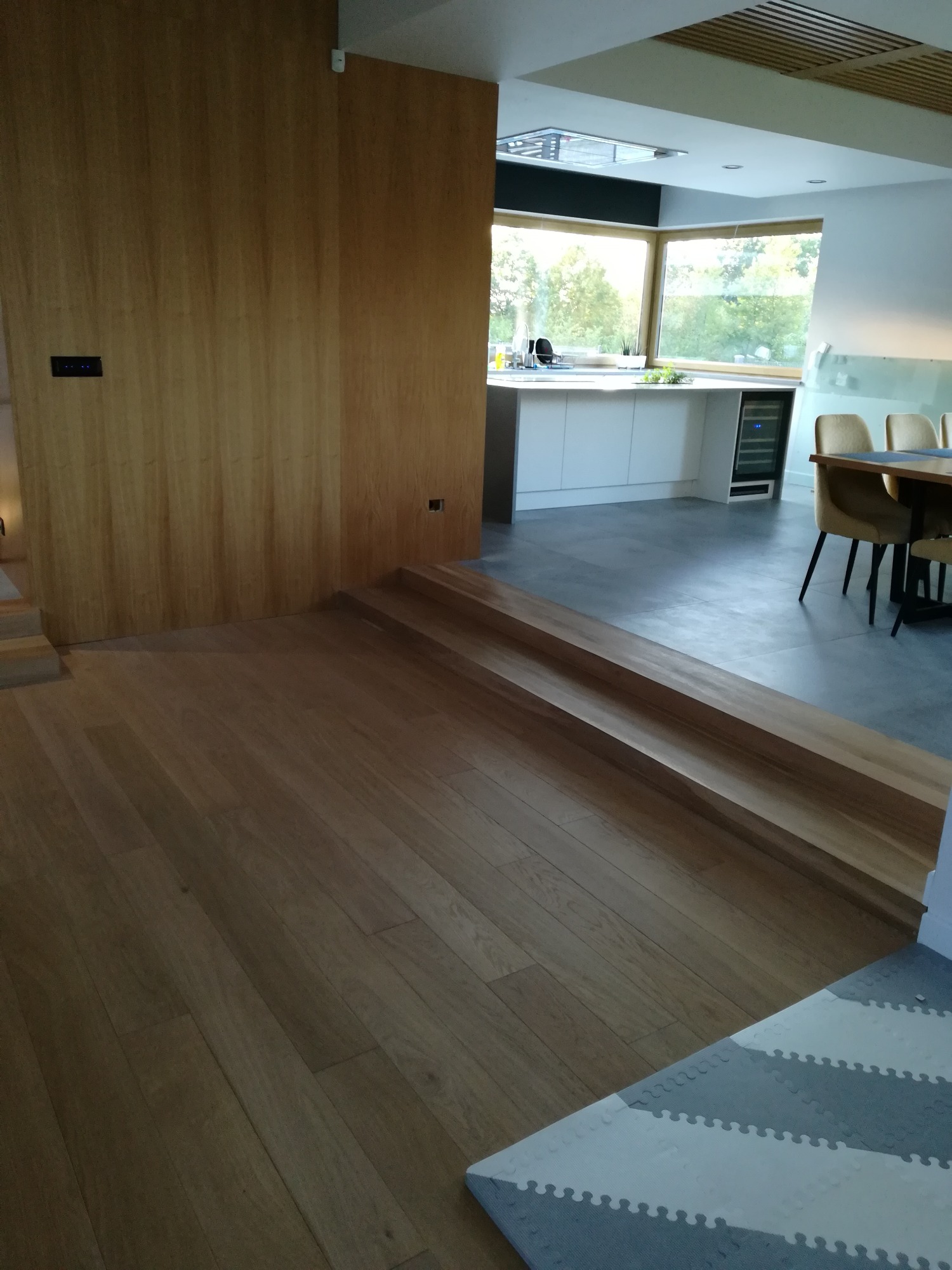 Jasna, drewniana podłoga przechodząca w schody w nowoczesnym wnętrzu z widokiem na kuchnię i jadalnię, minimalistyczny design.