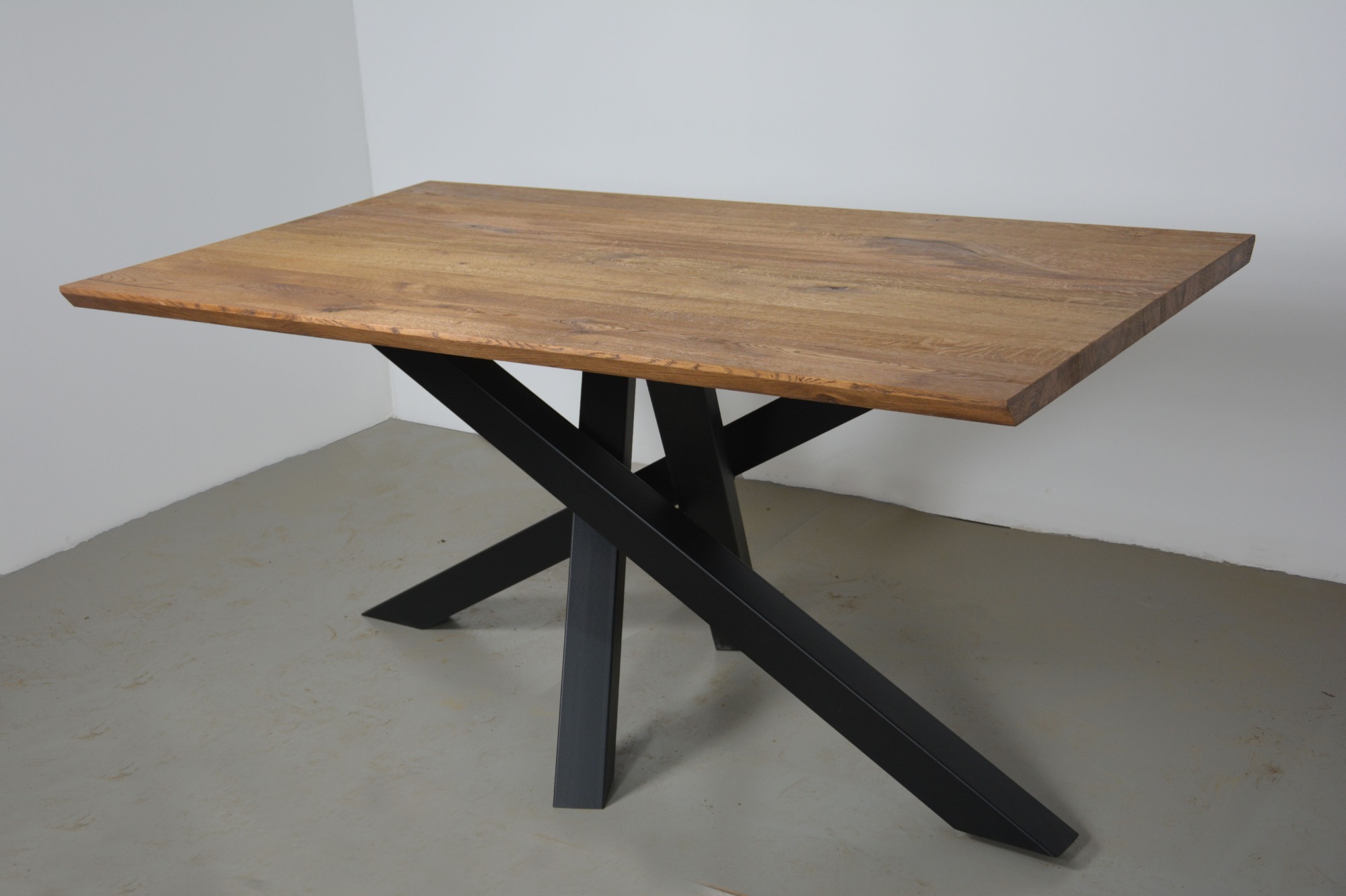 Prostokątny stół z blatem z jasnego drewna o widocznej strukturze słojów, wsparty na czarnych, metalowych nogach w kształcie litery X, na tle szarej podłogi i białej ściany.