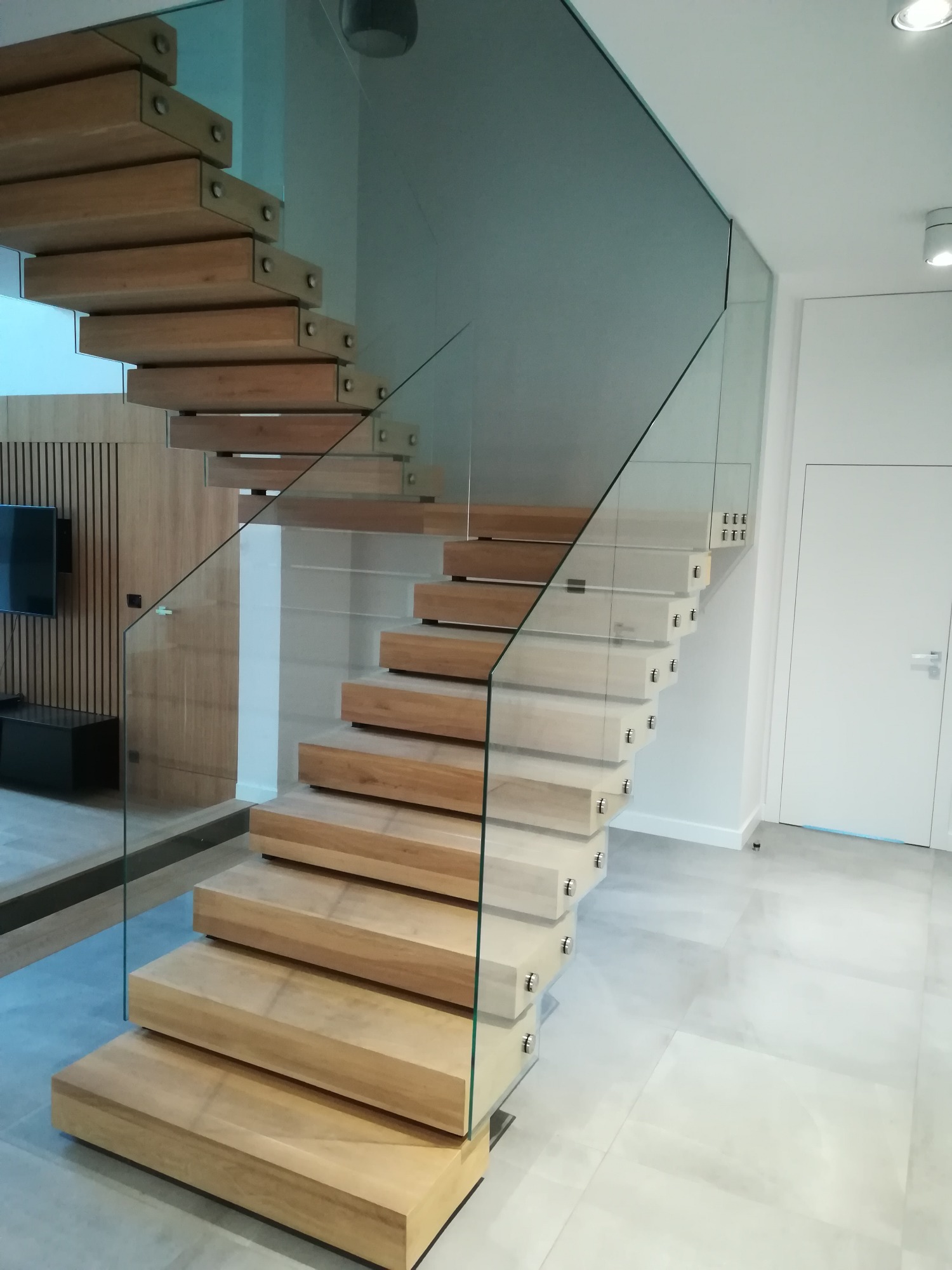 Nowoczesne, samonośne schody o drewnianych stopniach i szklanej balustradzie, zamocowanej za pomocą metalowych elementów, na tle ściany z drewnianymi panelami i telewizorem.