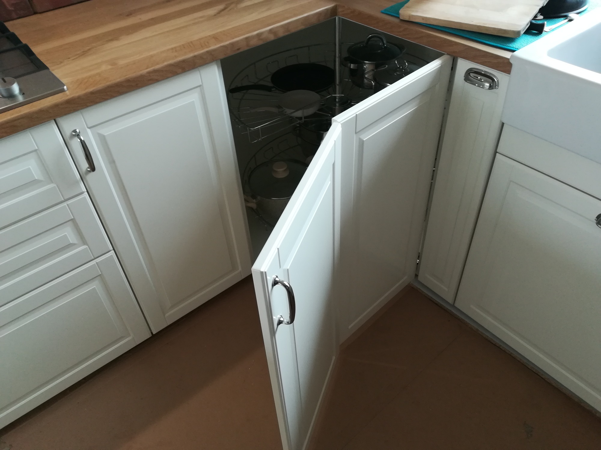 Otwarta szafka narożna w białej zabudowie kuchennej z obrotowym systemem przechowywania garnków i patelni, widoczny drewniany blat i zlew.