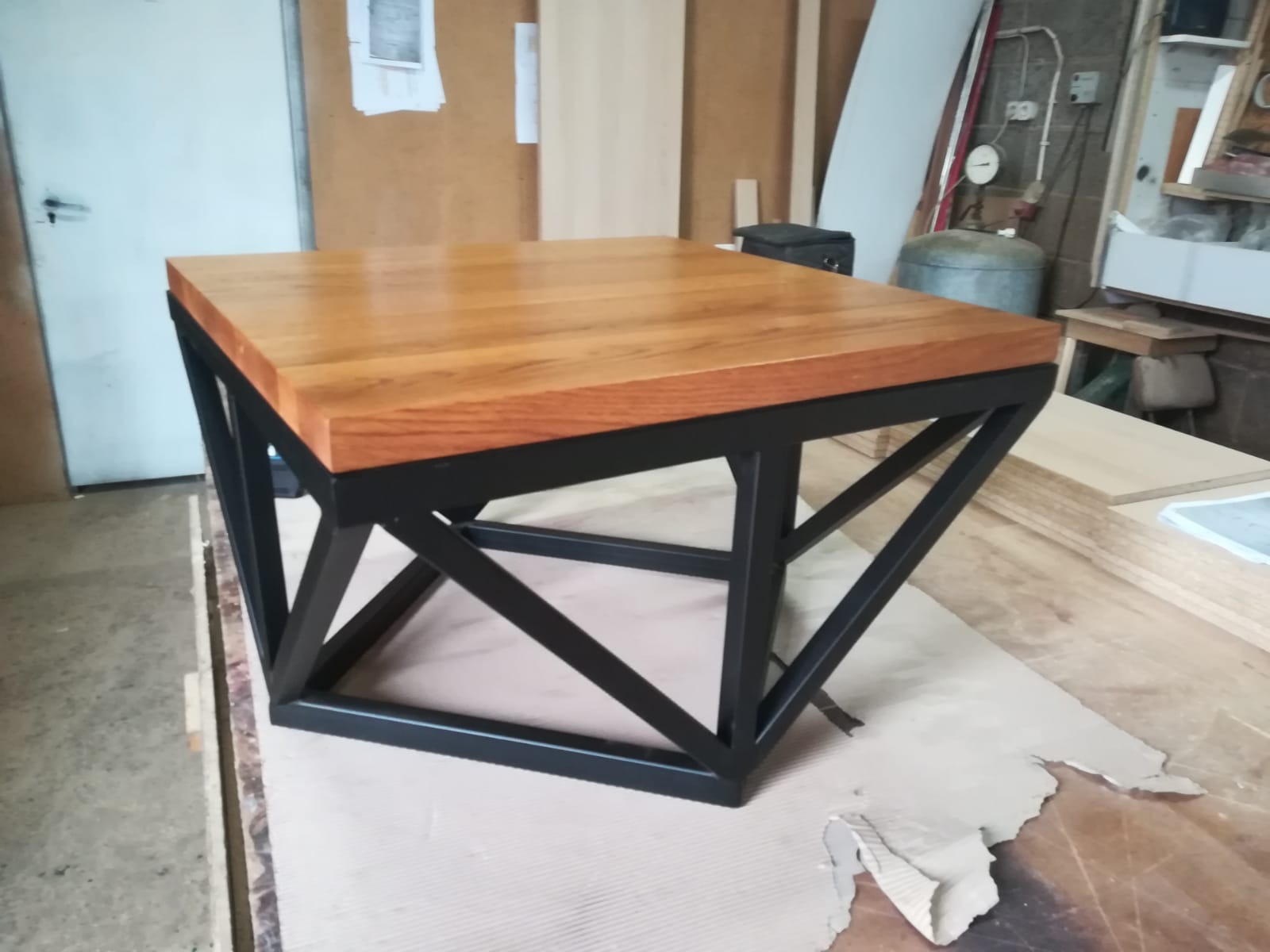 Kwadratowy stolik kawowy z blatem z jasnego drewna i czarną metalową podstawą o geometrycznym kształcie, widoczny w warsztacie stolarskim.