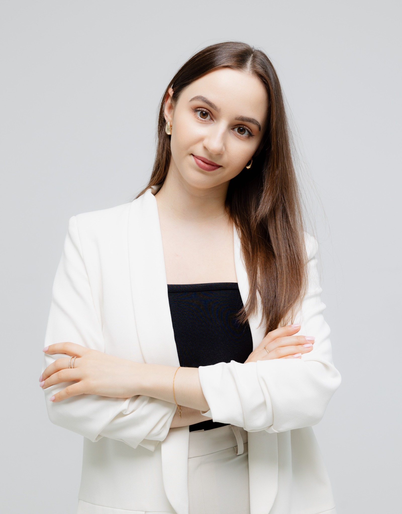 Portret kobiety w białym żakiecie i czarnym topie, z założonymi rękami, na jasnym tle. Widoczne złote kolczyki i pierścionek.