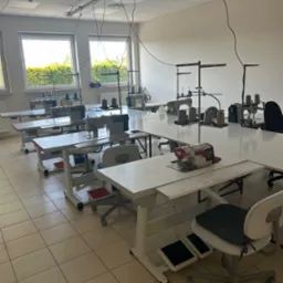 Jasne wnętrze szwalni z kilkoma stanowiskami pracy: maszyny do szycia, nici, krzesła. Widok na okna i zieleń na zewnątrz. Podłoga z płytek w odcieniach beżu.